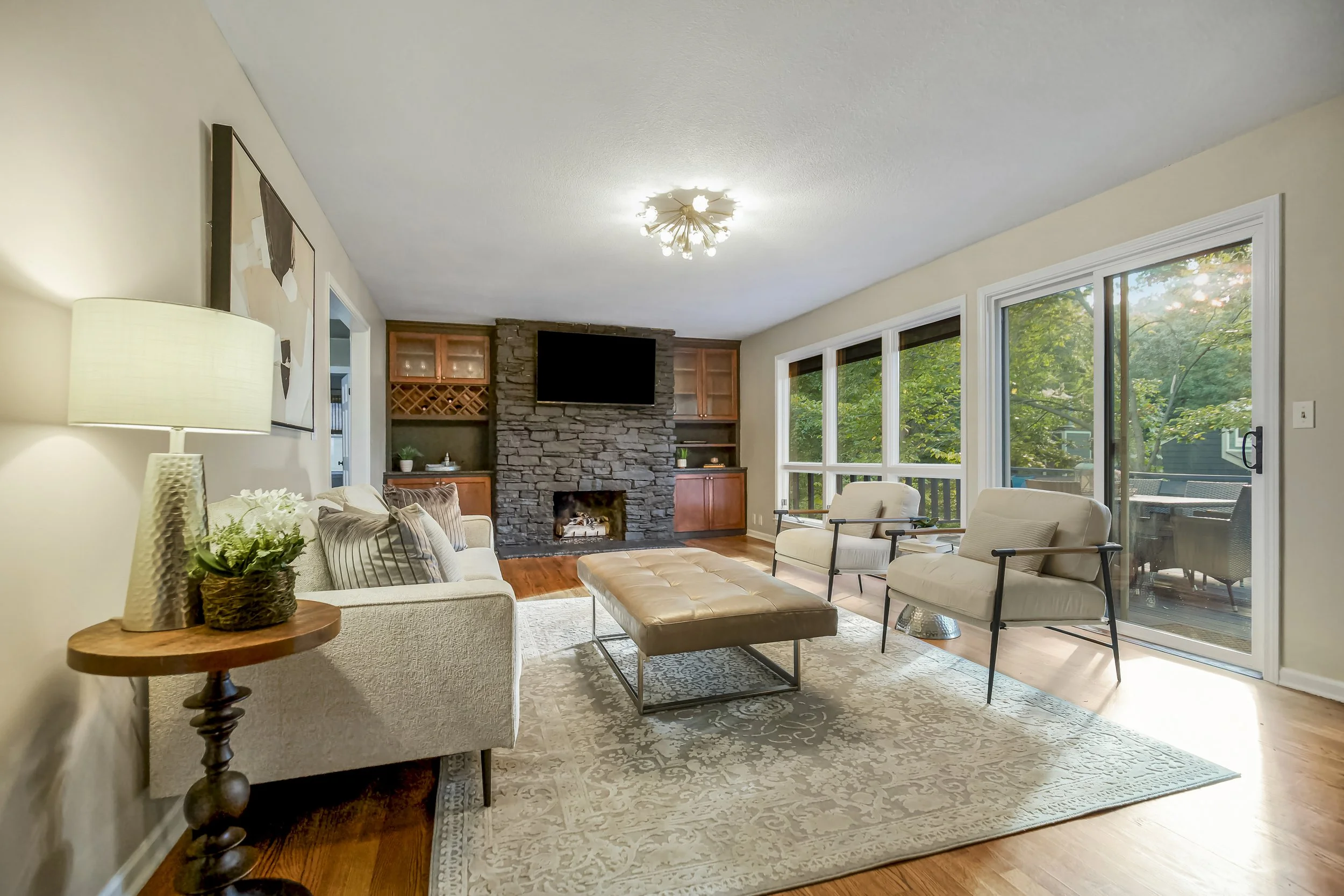Living room with large windows overlooking a patio, featuring a fireplace with a TV mounted above, a white sofa, two armchairs, a tufted ottoman, and a beige rug.