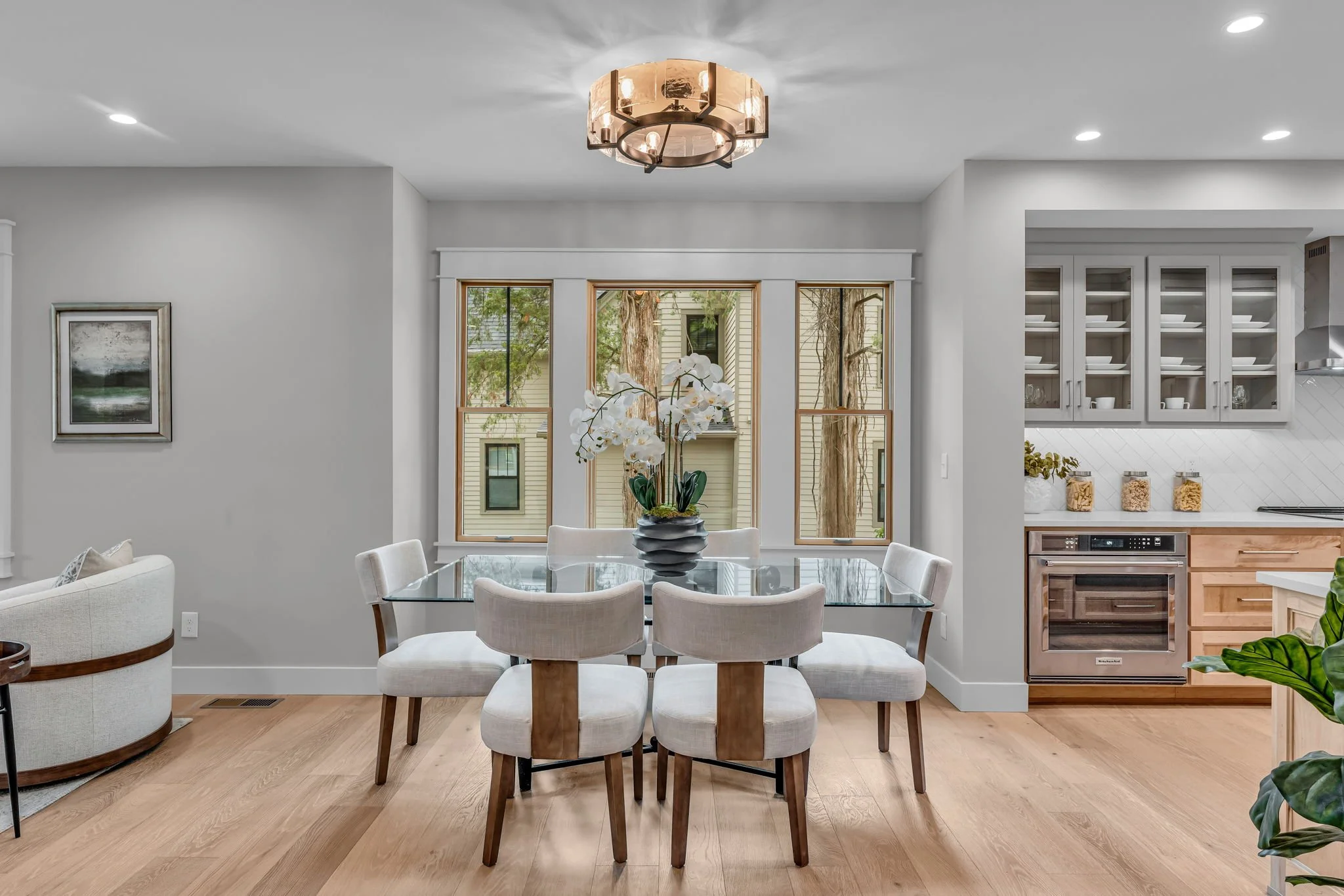 Dining area with a glass table, six white upholstered chairs, a floral centerpiece, large windows, light wood flooring, and an adjacent kitchen with white and wood cabinets.