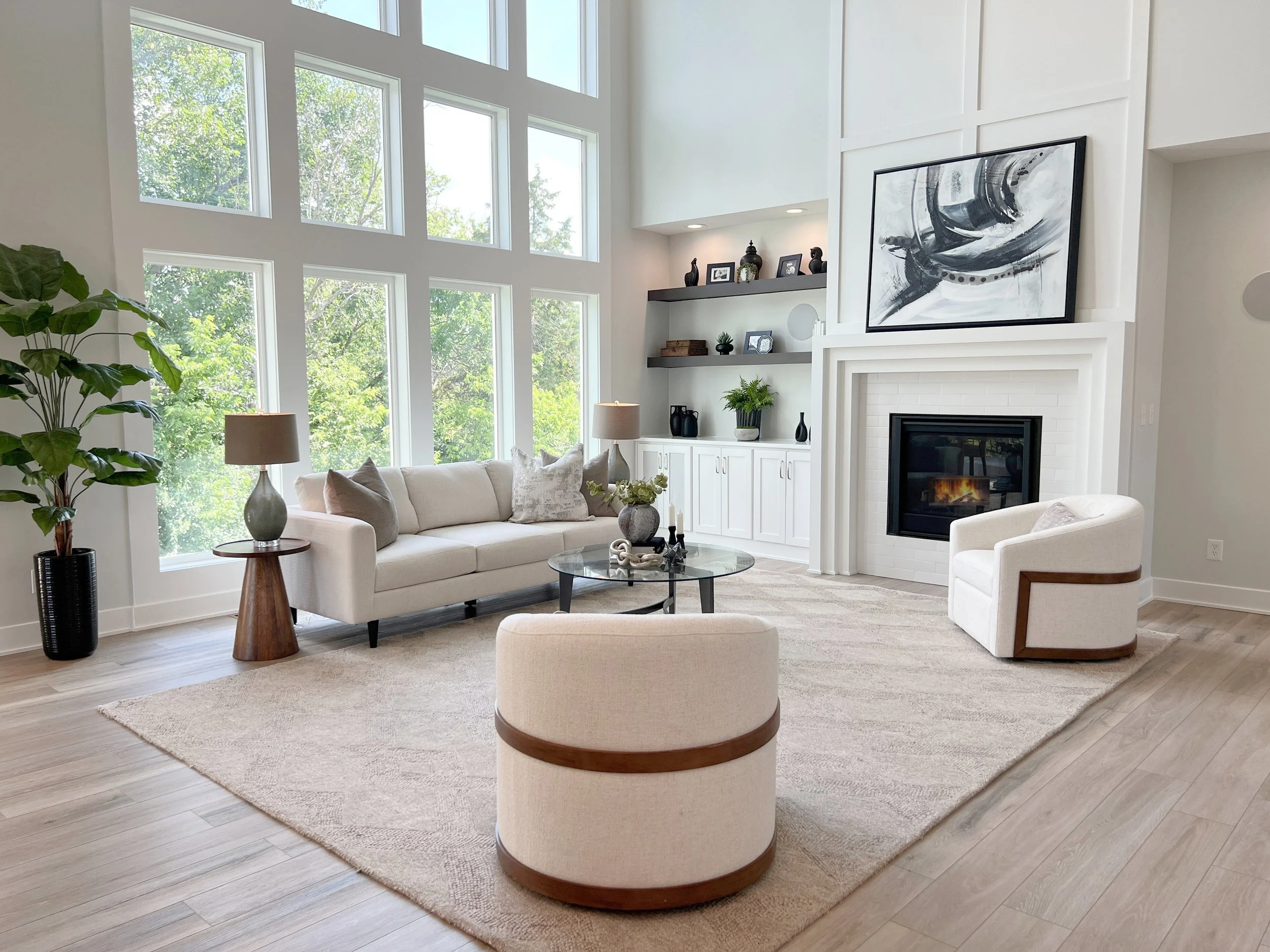 Bright living room with large windows, white sofa, two armchairs, a round glass coffee table, a fireplace, artwork, and potted plants.