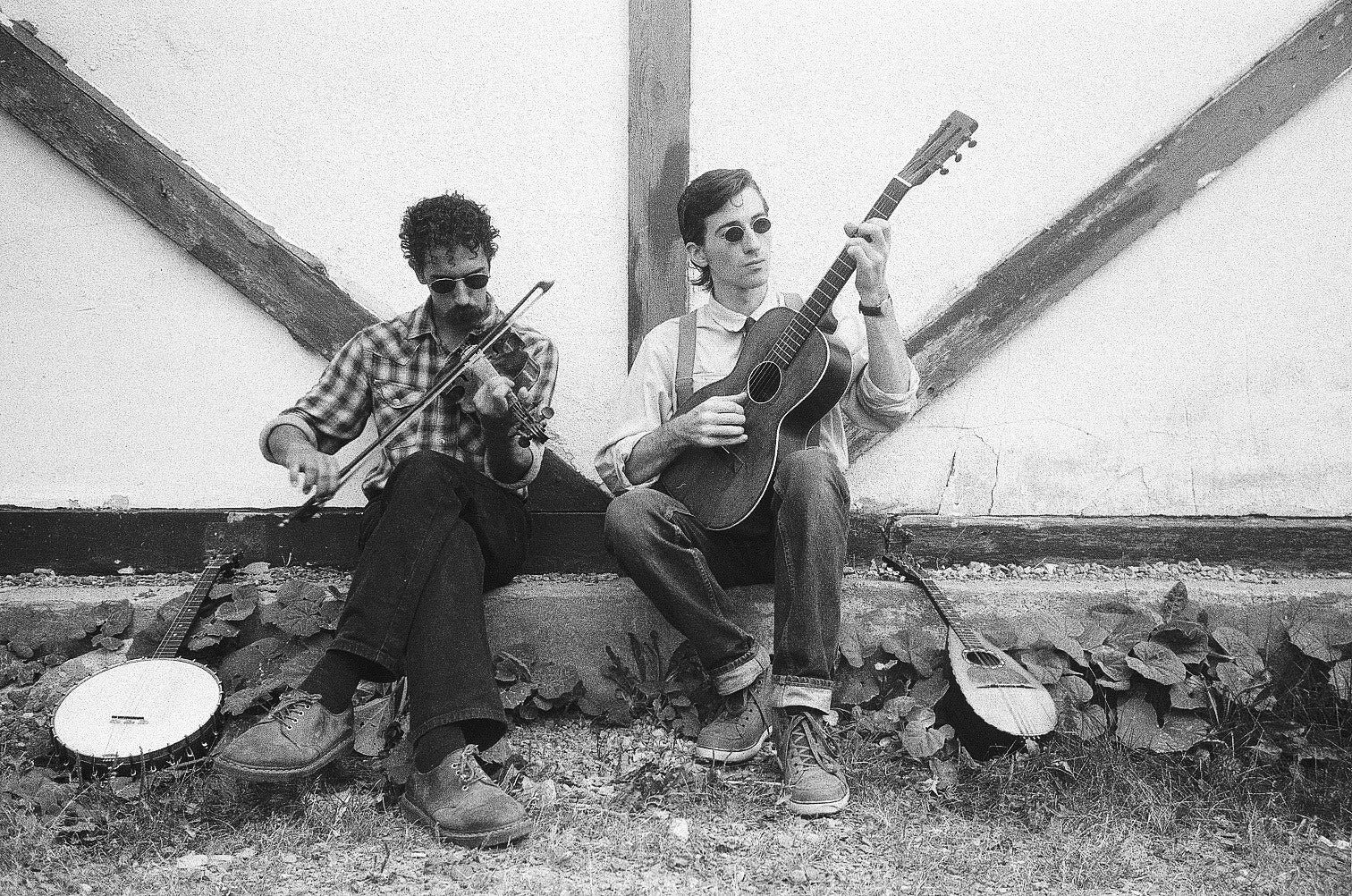 Two men sitting outdoors against a wall, playing musical instruments. One is playing a violin and the other is playing an acoustic guitar. They are both wearing sunglasses and casual clothing, and there are two additional guitars resting on the ground near them.