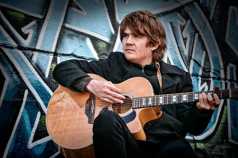 Man with brown hair playing an acoustic guitar in front of a graffiti wall.