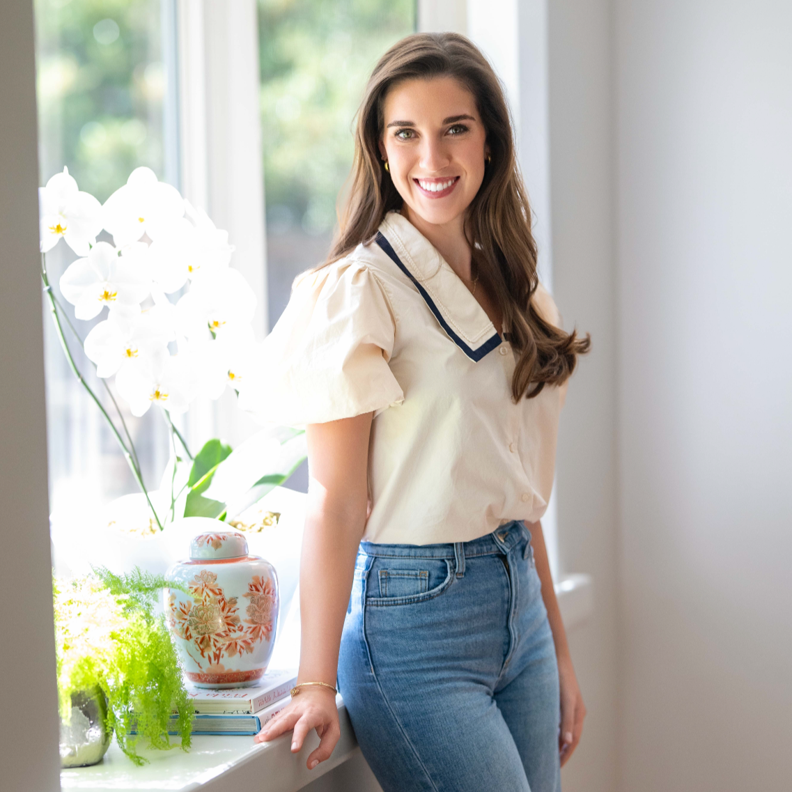 A young woman with long brown hair, wearing a cream-colored blouse with navy trim and blue jeans, smiling and standing by a window with a potted orchid, a decorative vase, and books on a windowsill.