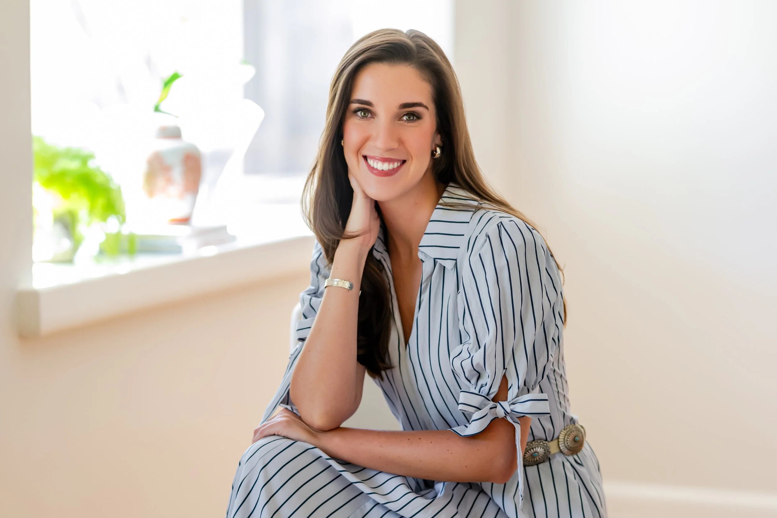 A woman with long brown hair and green eyes smiling, seated indoors near a window with potted plants.