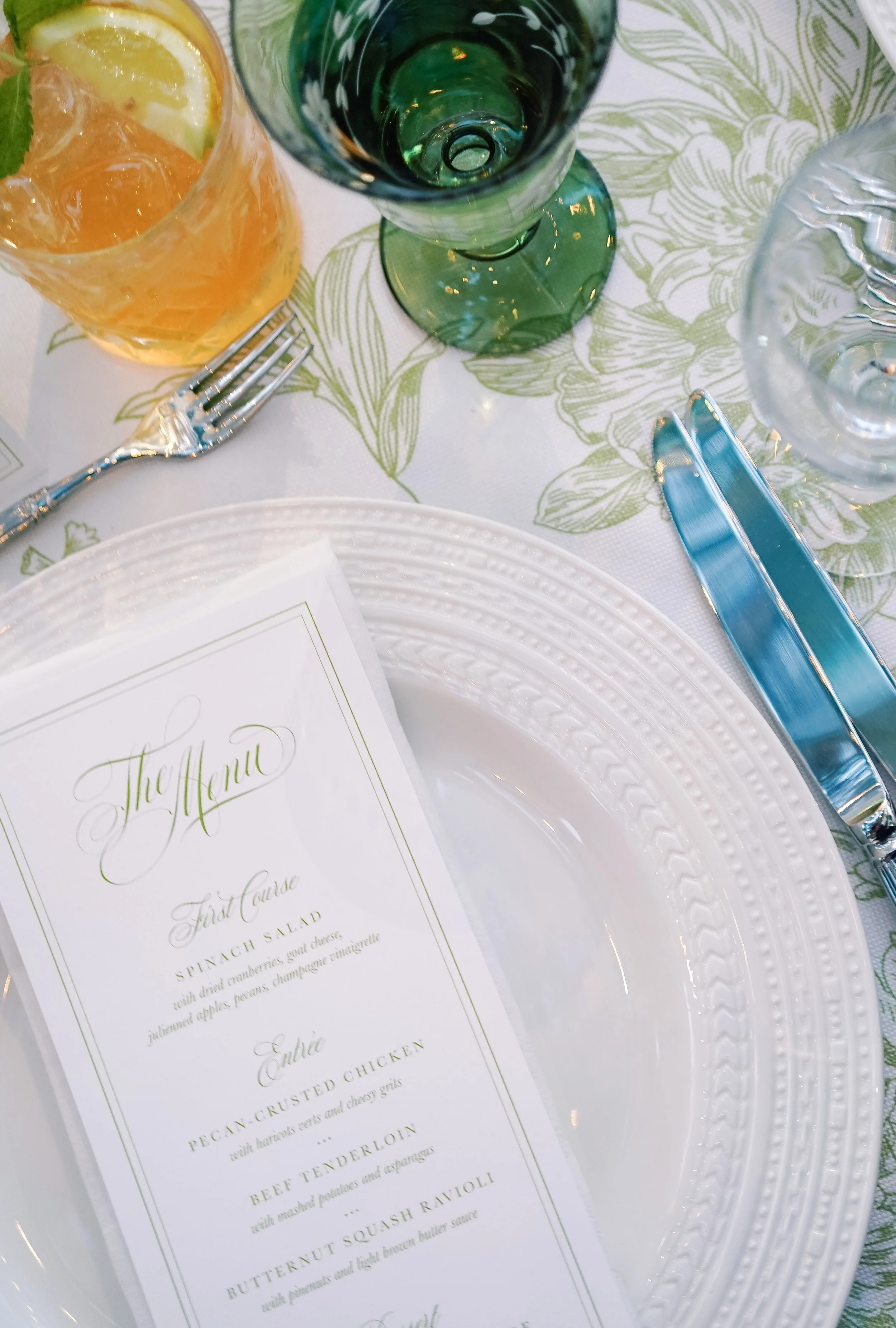 A place setting at a formal dining table with a white decorative plate, a menu, a fork, a knife, and a spoon, with drinking glasses including a green wine glass, a clear glass with ice and lemon, and a water glass, on a tablecloth with a green floral