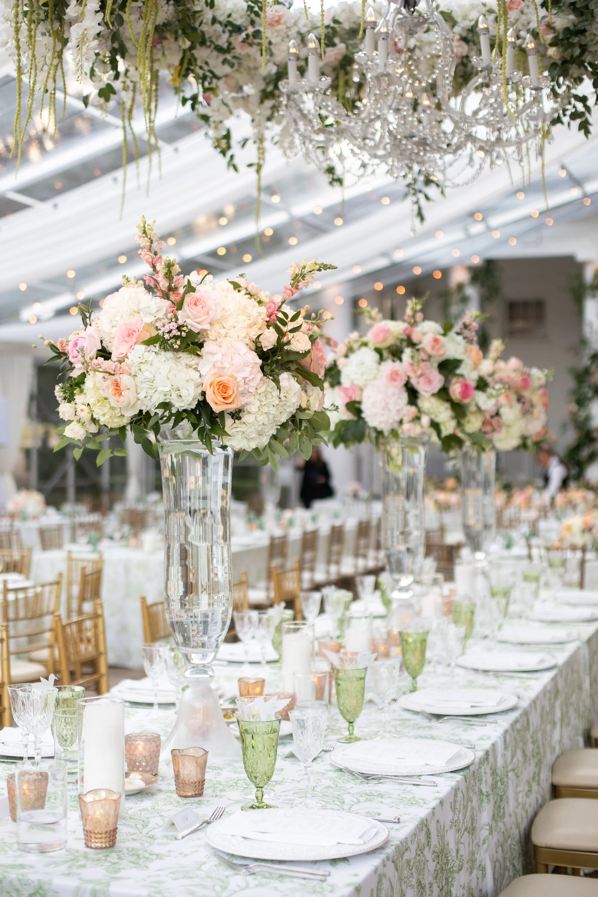 Elegant wedding reception table decorated with tall floral centerpieces of pink and white roses and hydrangeas in glass vases, with candles and transparent tableware.