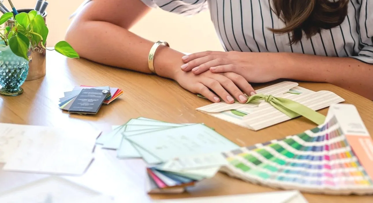 Person working on a craft project at a table with paint swatches, fabric samples, and color cards, wrapping a gift with green ribbon.