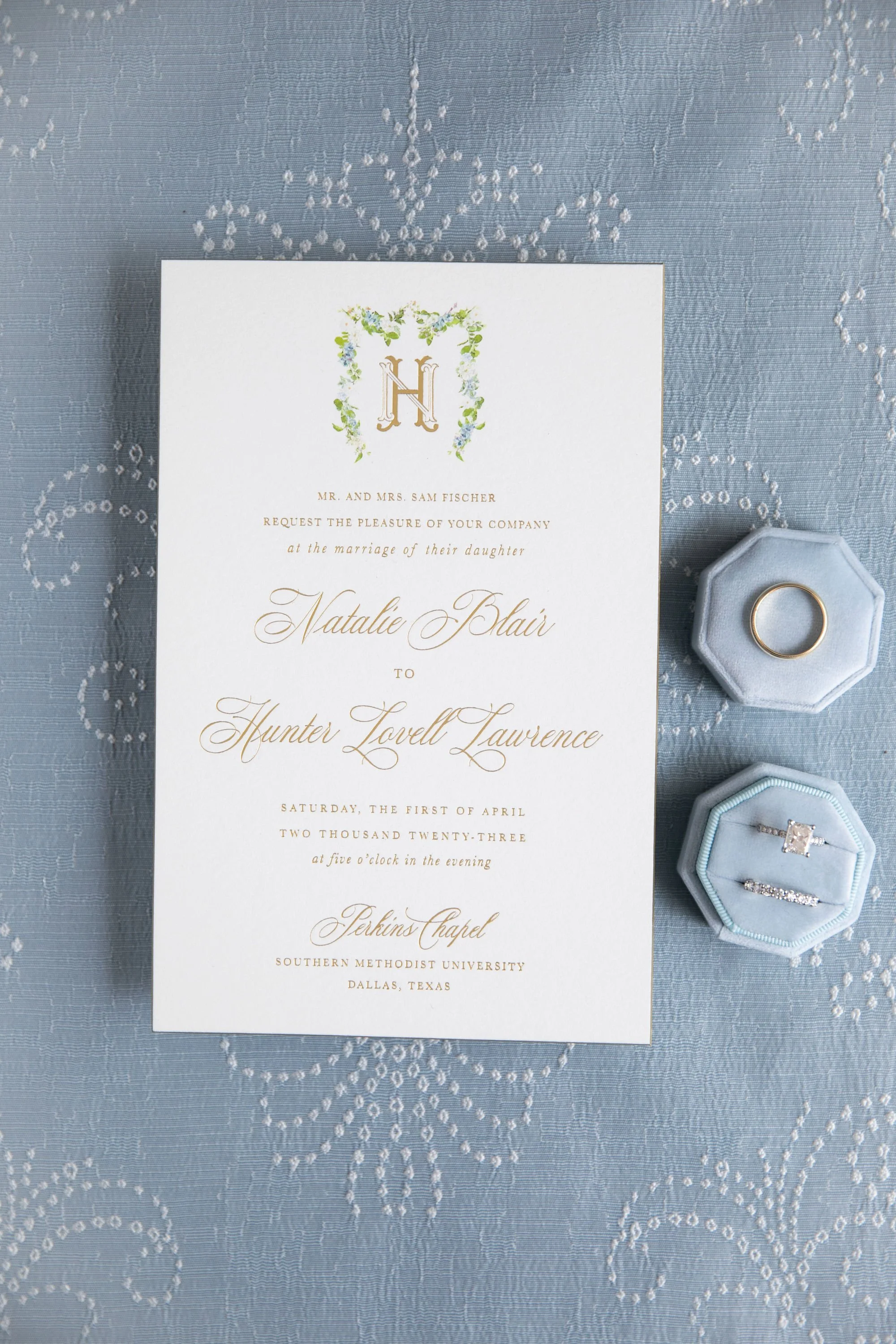 Wedding invitation on a blue embroidered fabric with two rings in a light blue ring box.