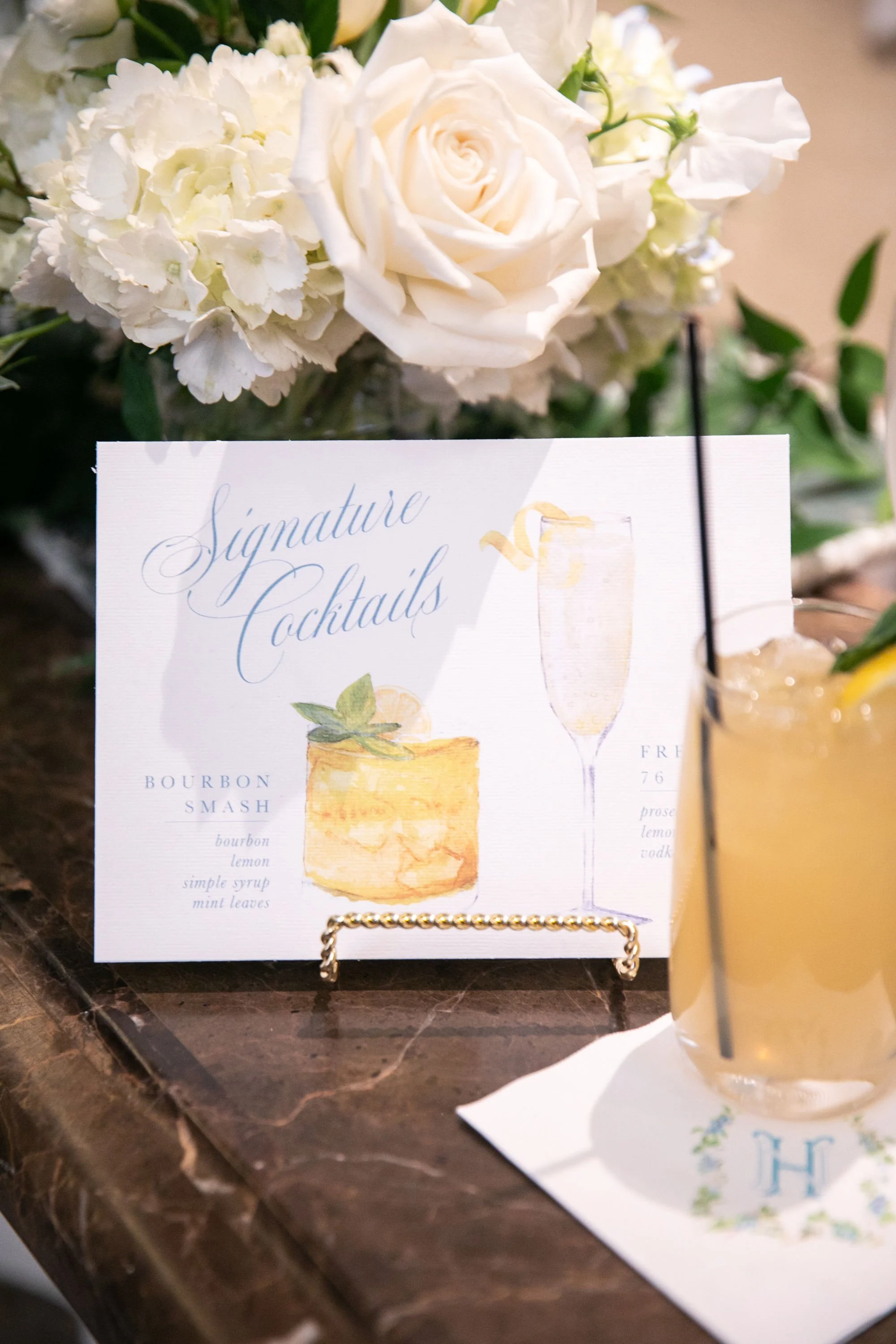 A sign displaying two illustrated cocktails labeled 'Signature Cocktails,' with a bouquet of white roses and hydrangeas behind it and a glass of yellow cocktail with a lemon wedge in front.