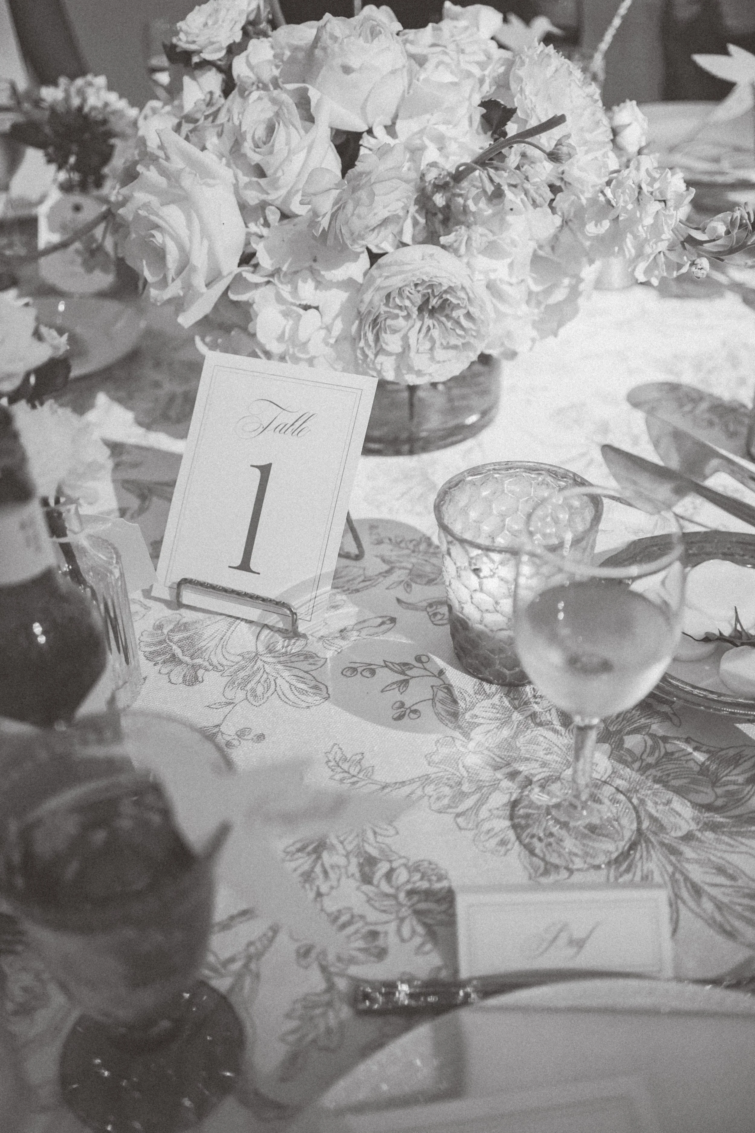 Black and white photograph of a formal dinner table setup with a large floral centerpiece, a table number card labeled "Table 1," glassware, cutlery, and a patterned tablecloth.