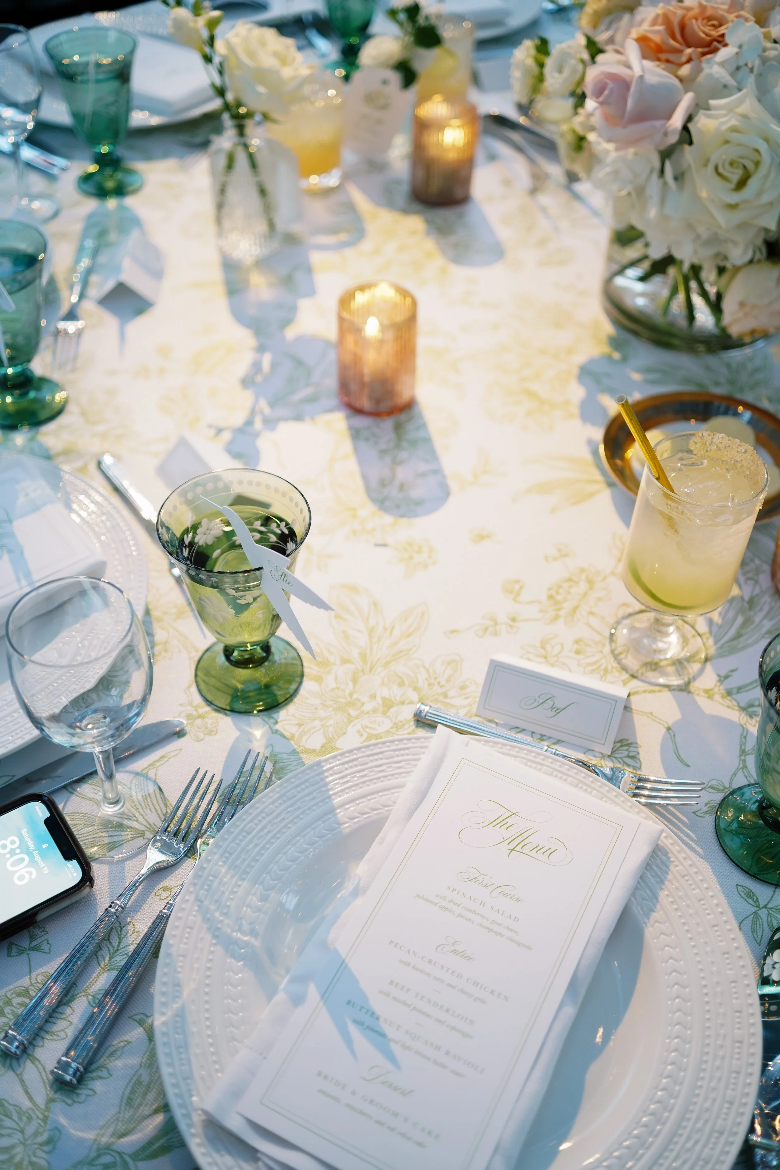 A beautifully set wedding banquet table with floral centerpiece, candles, wine glasses, and a printed menu. The table features elegant glassware and cutlery, with a small place card and a cocktail drink.