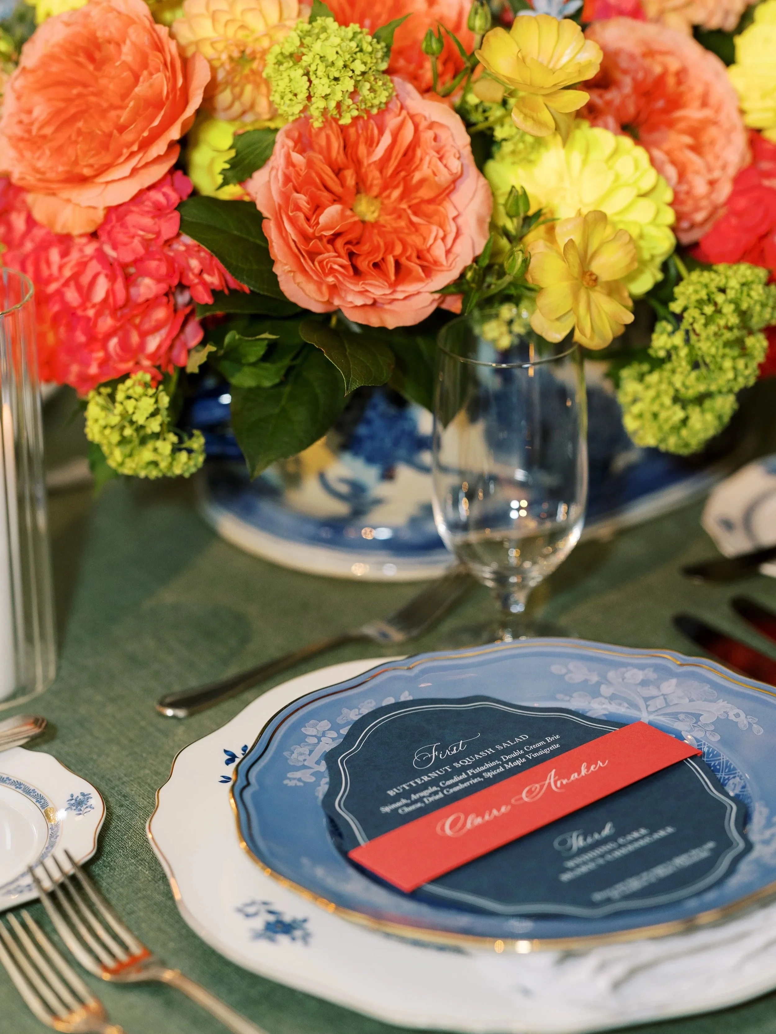Elegant dining table with a floral centerpiece of pink and yellow flowers, fine china plates with gold trim, silverware, an empty wine glass, and a menu card.