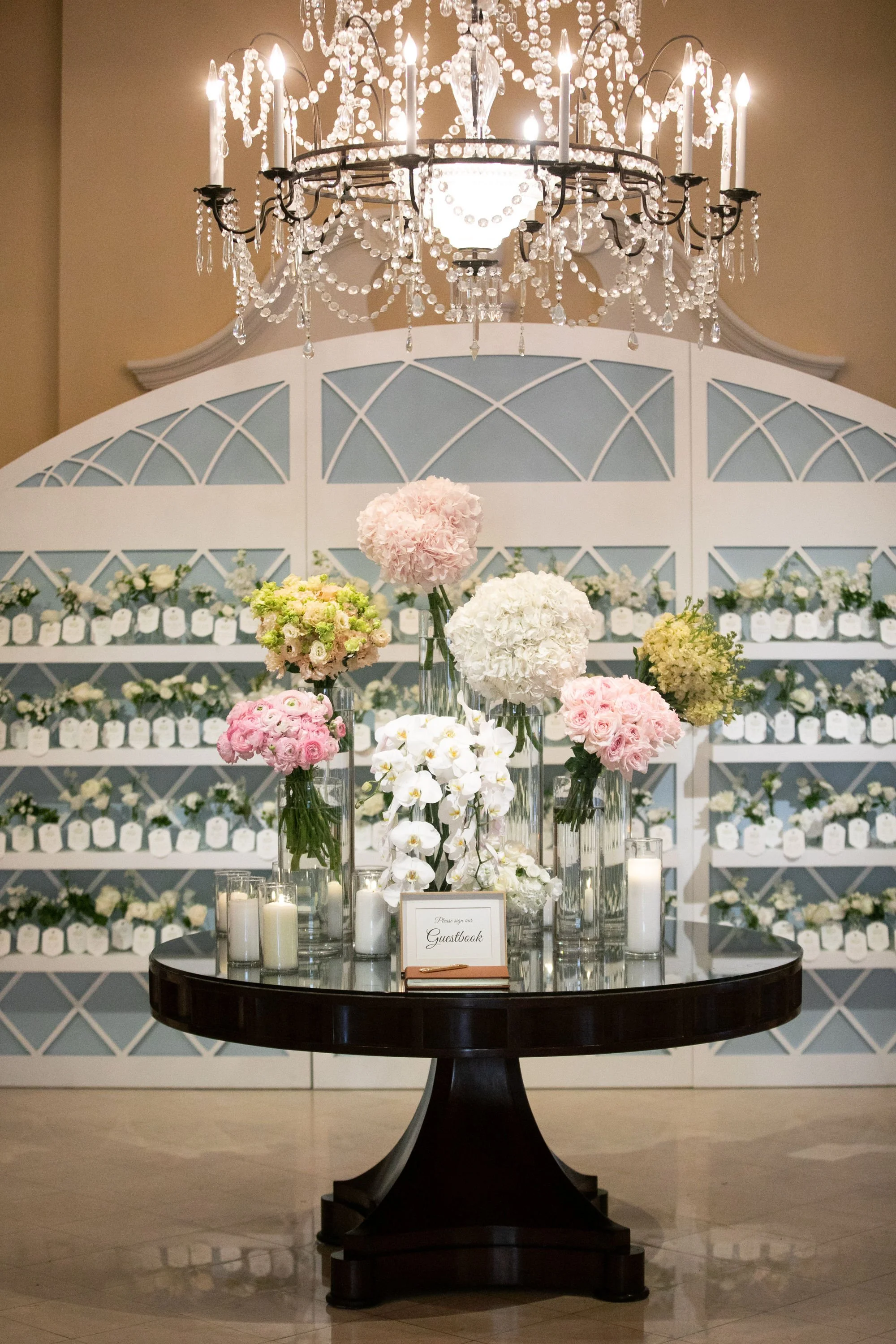 Elegant guestbook display with pink, white, and green floral arrangements in tall vases, surrounded by white candles on a dark wooden table, set against a backdrop of shelves filled with white decorative jars and topped with a crystal chandelier.
