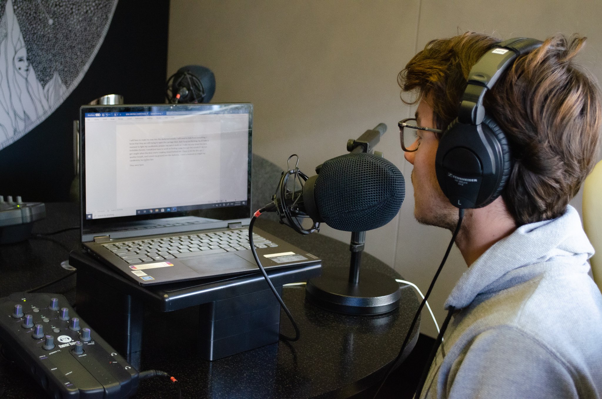A teenage boy with brown hair, sitting in front of a laptop and speaking into a microphone
