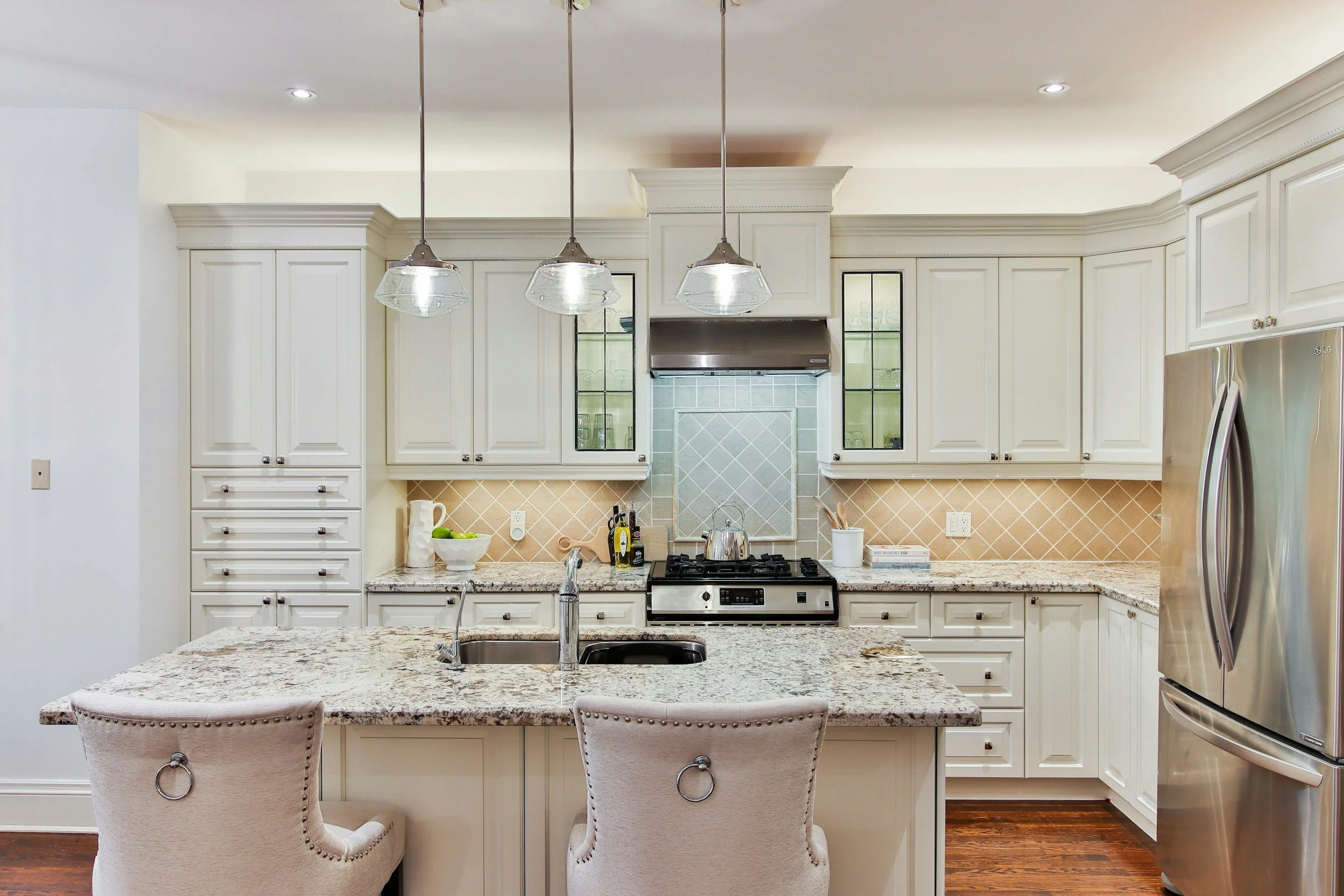 Kitchen with white cabinets, granite countertops, stainless steel appliances, and two beige upholstered chairs at the island. Lighting fixtures hang above and the backsplash features beige tile.