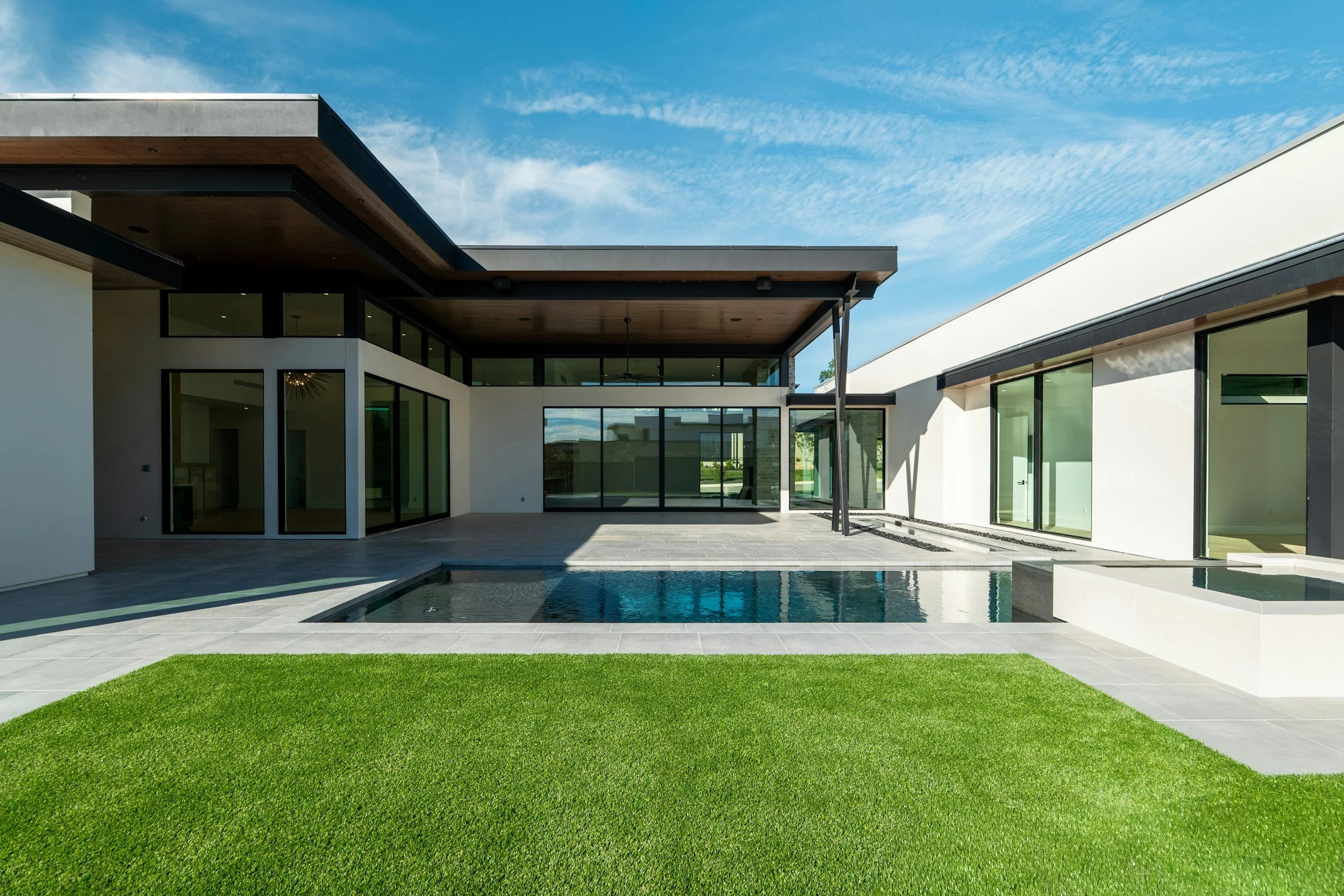 Modern house with large glass windows, swimming pool, and green lawn under a blue sky with scattered clouds.