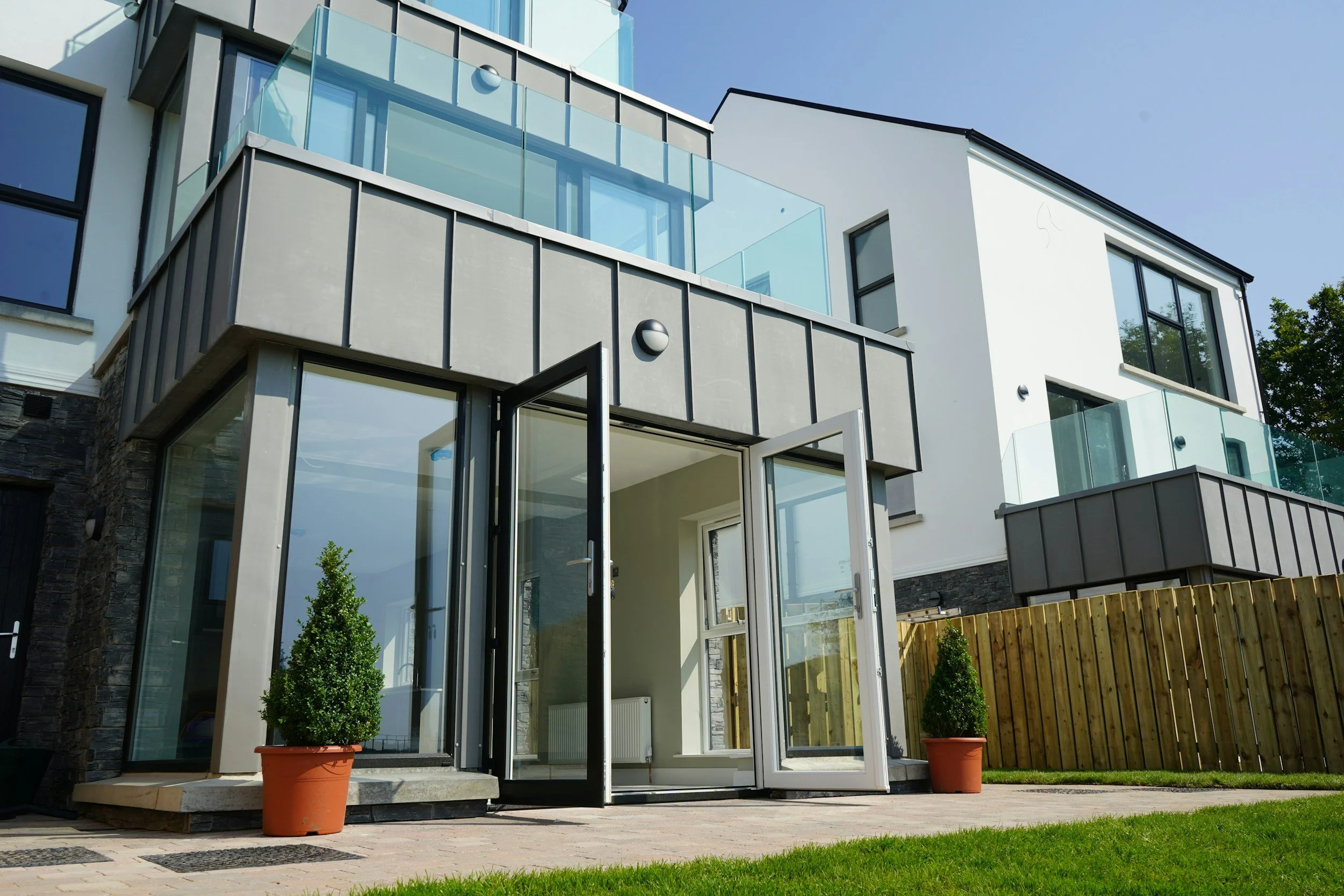 Modern white multi-story building with glass balconies and large windows, potted plants, and a wooden fence. The sky is clear and blue.