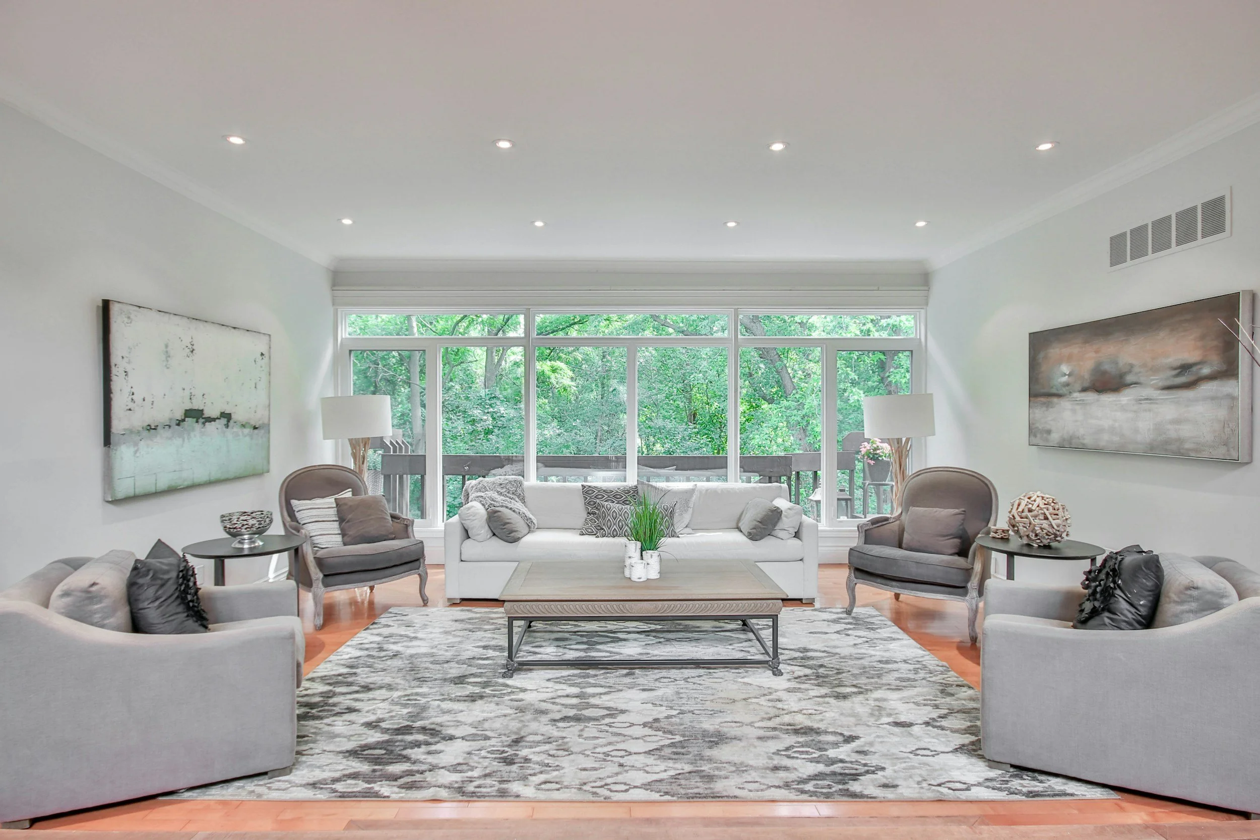 Bright living room with large windows overlooking trees, featuring white couches, gray armchairs, modern art, and a patterned rug.