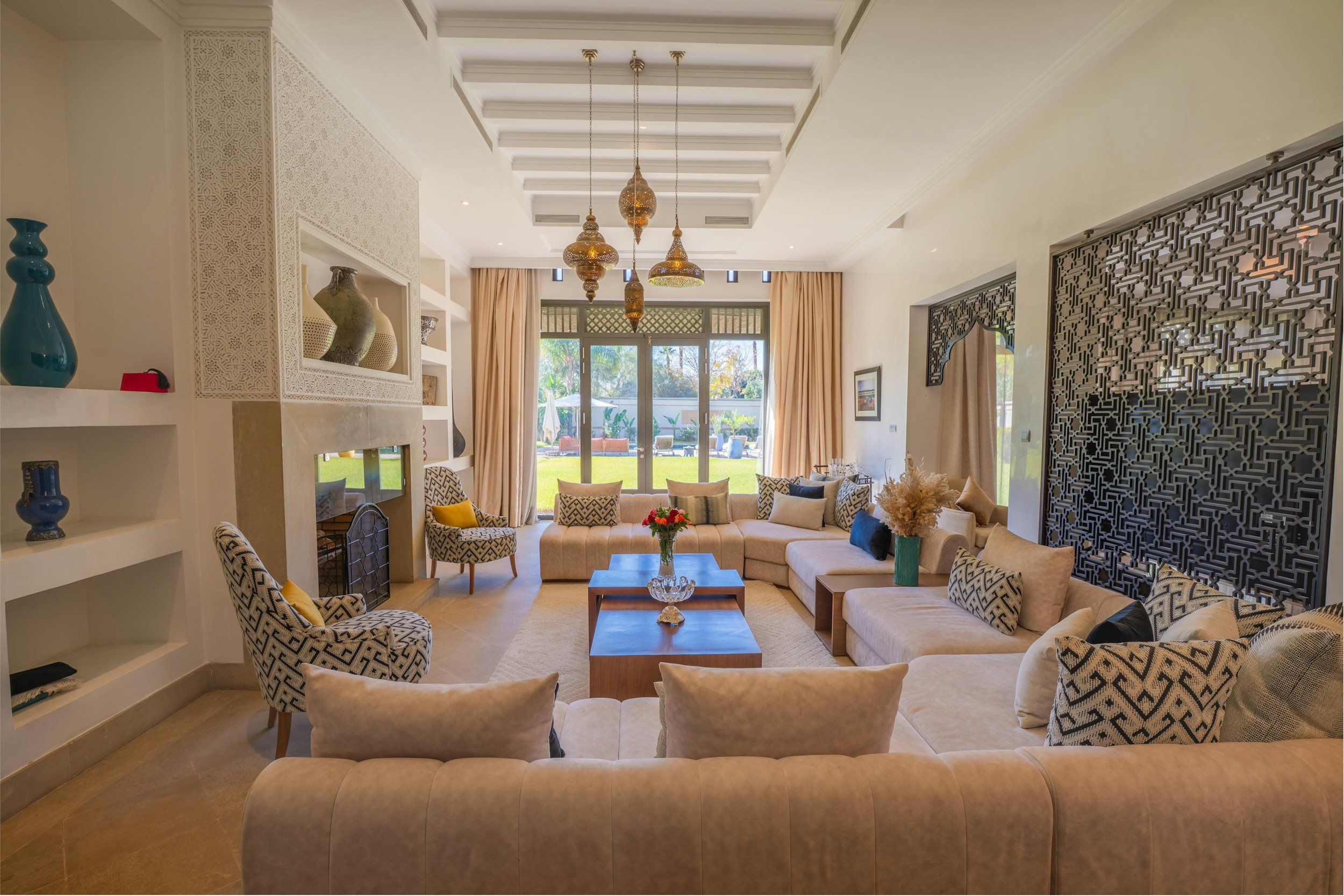 A spacious living room with a high ceiling featuring multiple hanging pendant lights. The room has large windows with beige curtains, allowing natural light to illuminate the space. It is furnished with multiple beige and patterned armchairs and a sectional sofa, all arranged around a central wooden coffee table with a floral arrangement. Decorative vases and art pieces are displayed on built-in shelves and on the wall, with a view of a garden visible through the windows.