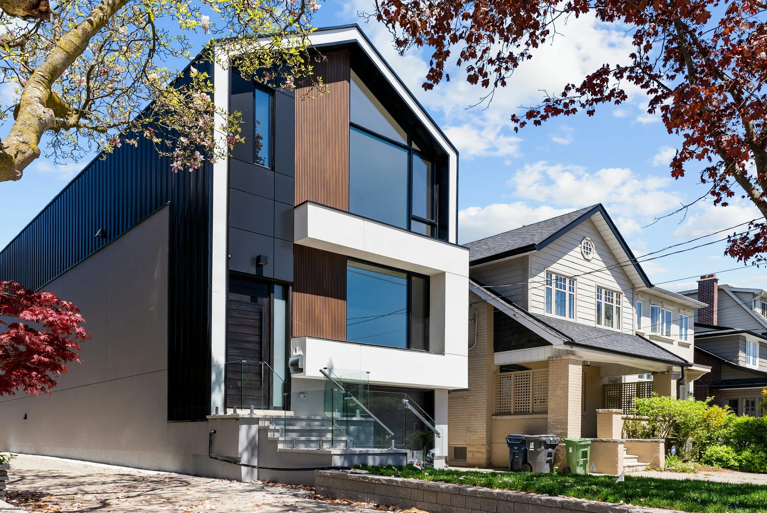 Modern multi-story house with large windows, dark exterior siding, and a staircase leading to a balcony, surrounded by trees with green and red leaves and a clear blue sky.