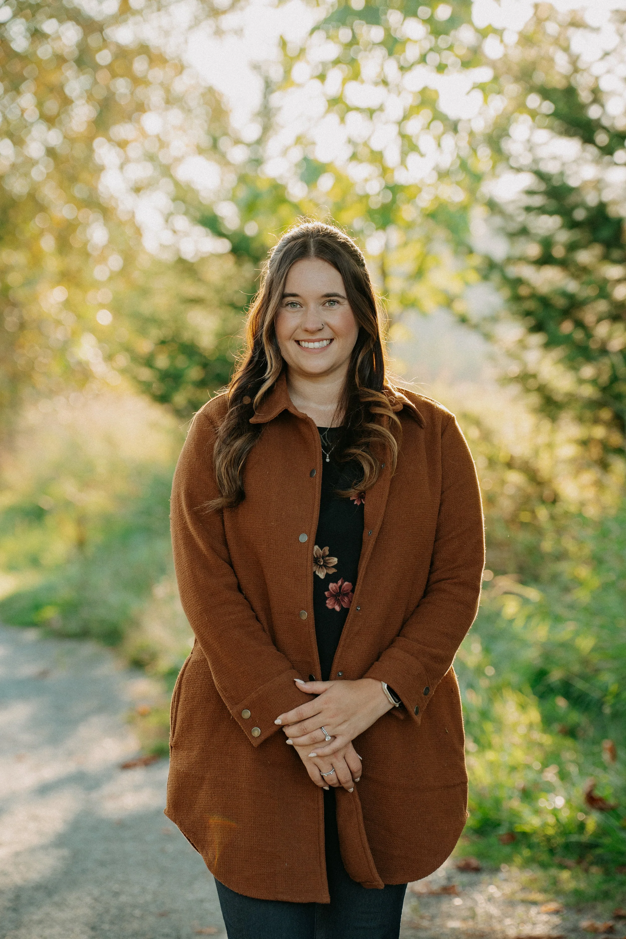 A woman with long brown hair smiling outdoors on a dirt path surrounded by trees with green and yellow leaves during fall.