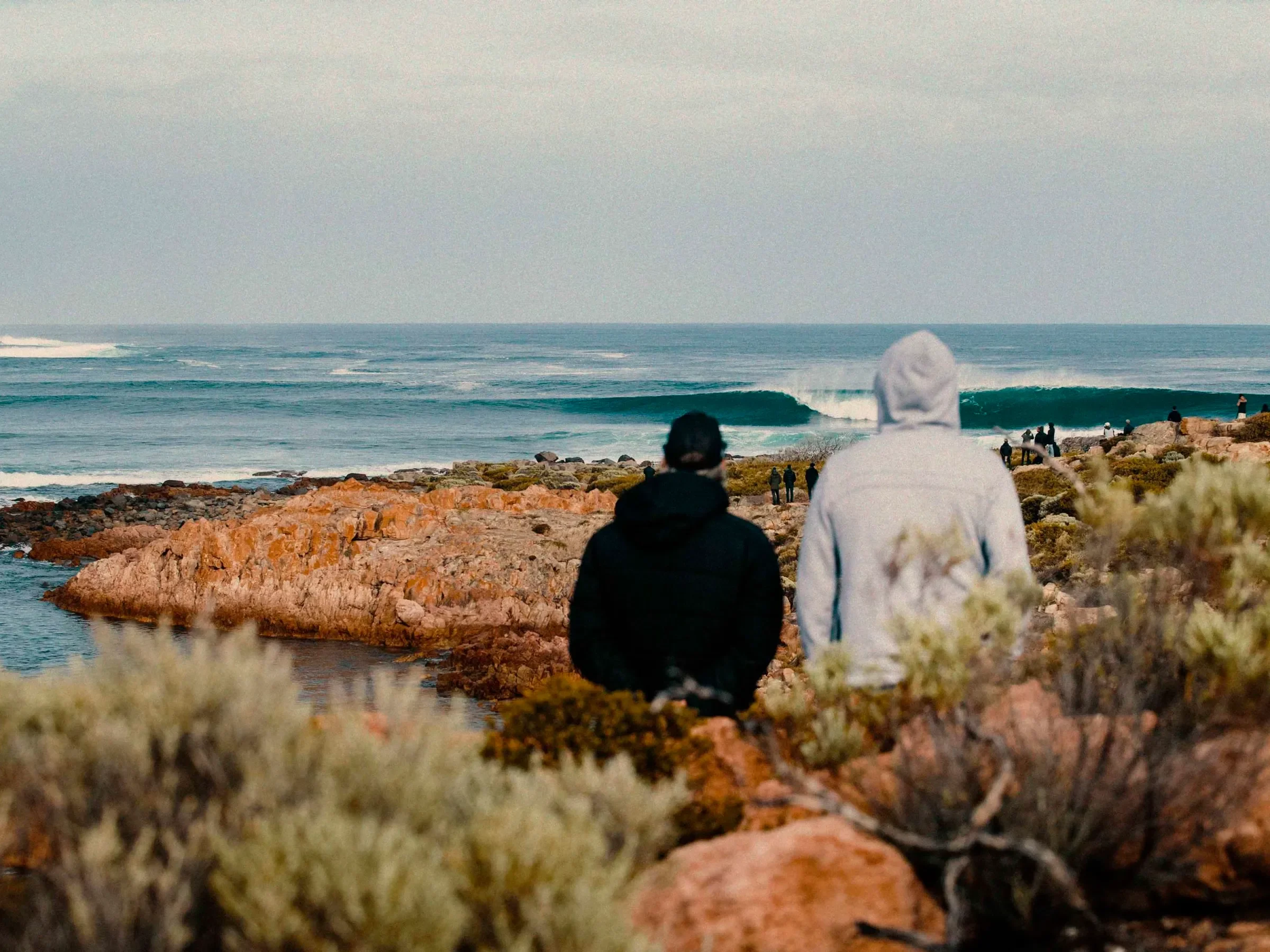 Two people walking along a rocky shoreline with a view of the ocean and Yallingup waves in the background. The Timothee Resort. Retro Motel Accommodation by the Beach in Busselton, WA. Boutique Holiday Getaway at Margaret River Hotel.