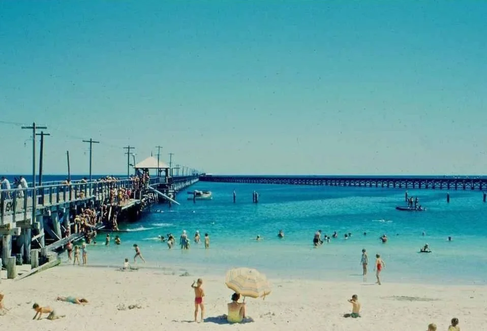 A Vintage 1980s postcard scene of people enjoy a day at the beach with the Busselton Jetty into the ocean, some swimming in the water, others sunbathing or standing on the sand, under a clear blue sky. The Timothee Resort. Retro Motel Accommodation