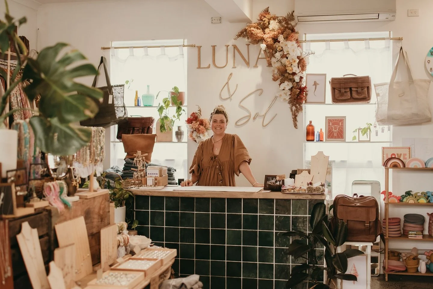 A smiling woman standing behind a boutique store counter decorated with flowers, plants, and various merchandise, with a sign in the background reading 'LUNA & Sol'.