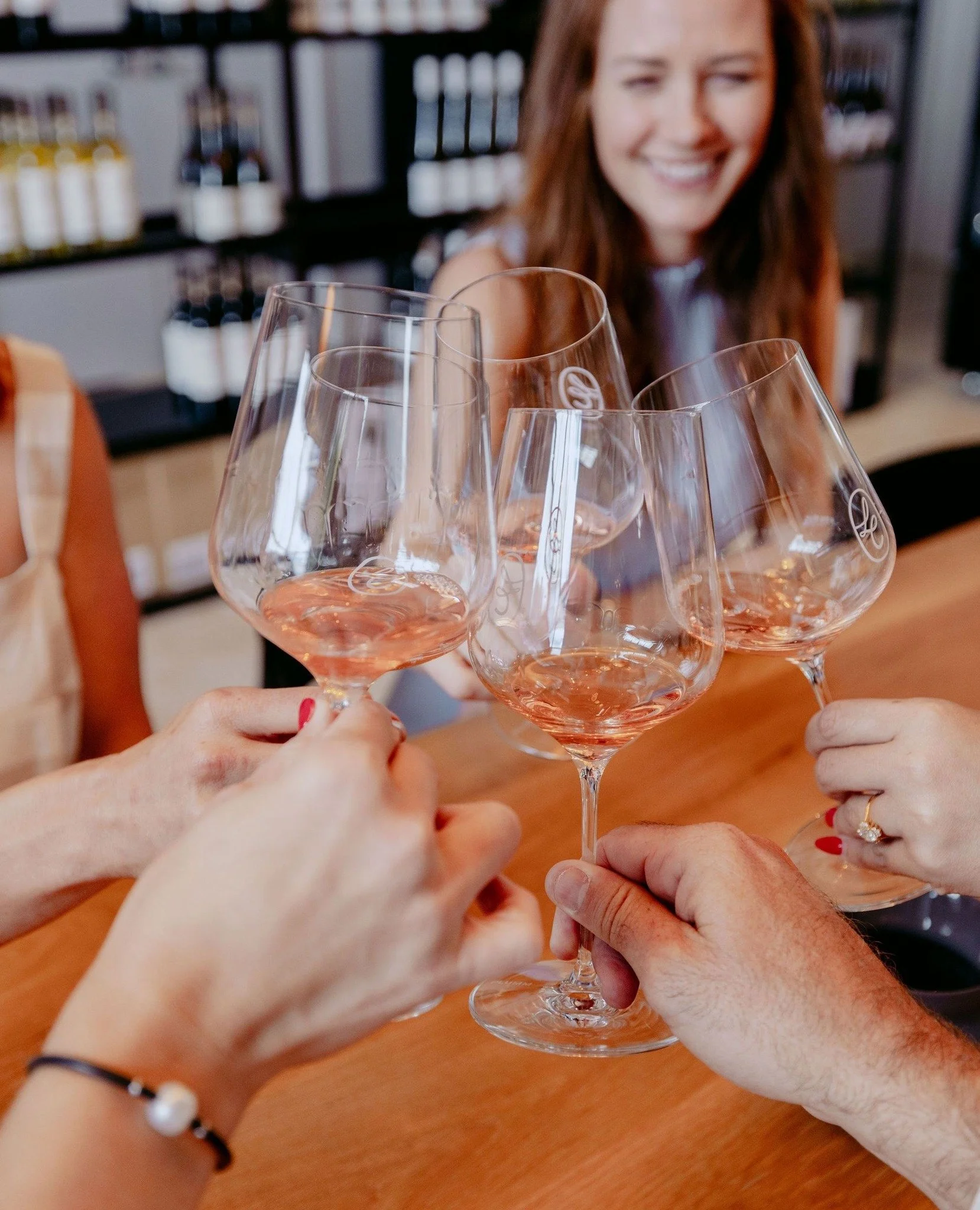 A group of people are toasting with glasses of rosé wine in close-up, with a woman smiling in the background at a wine shop or tasting room.