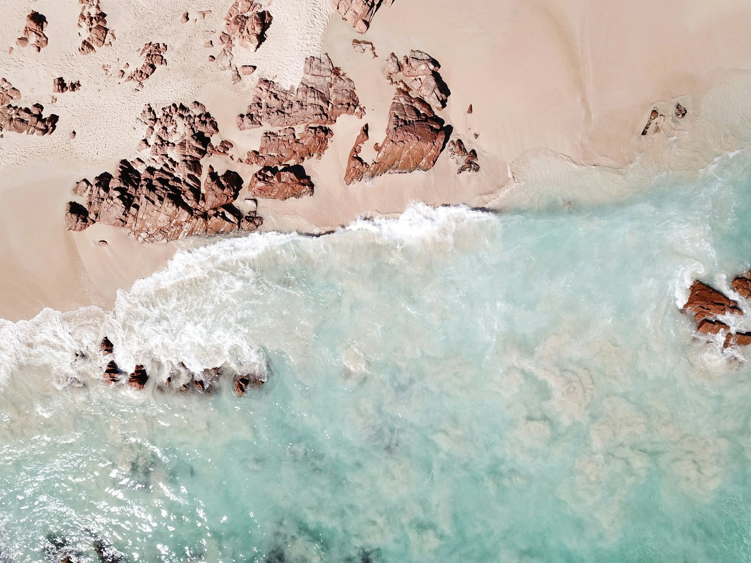 Aerial view of a sandy beach with reddish-brown rocks and ocean waves crashing onto the shore.