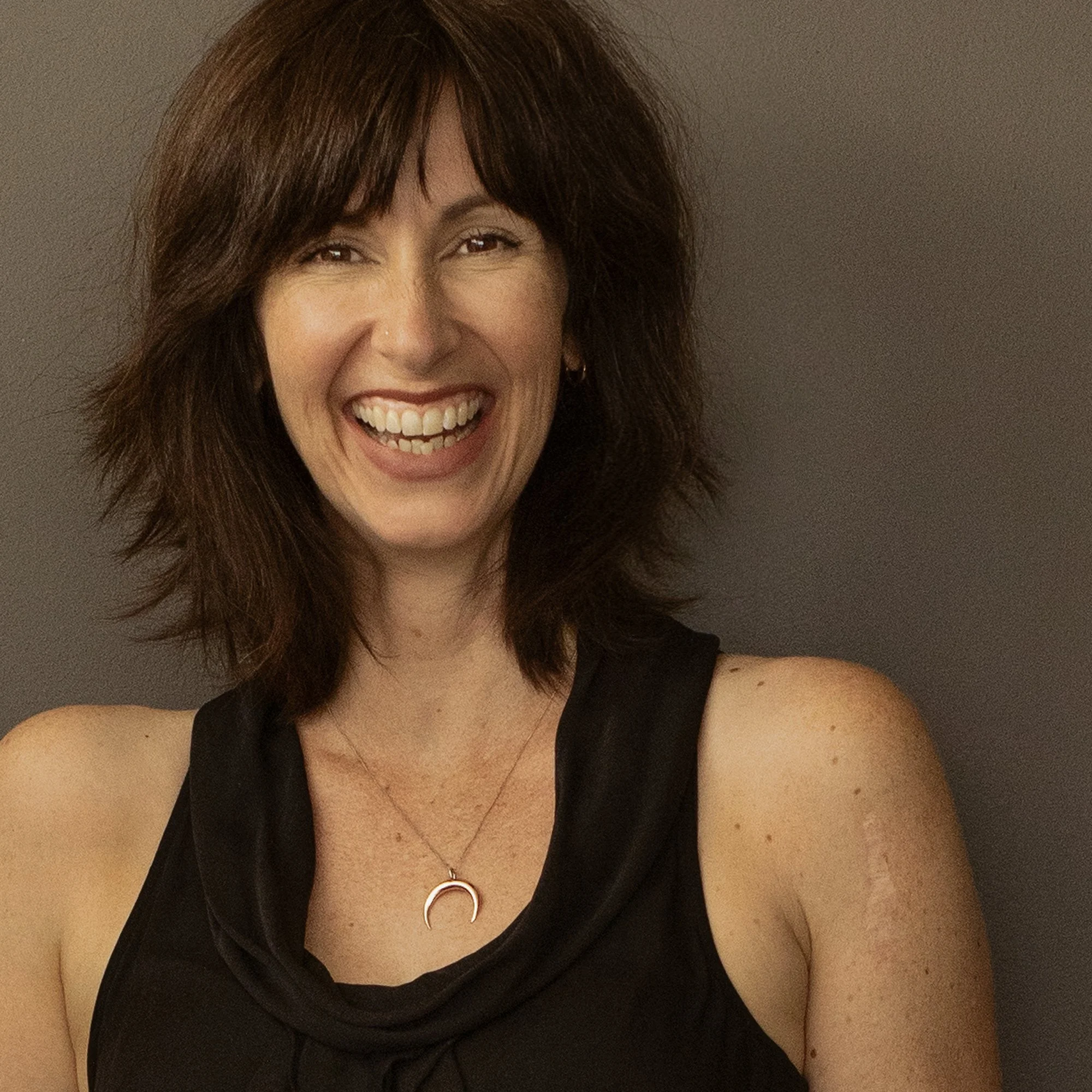 Human Design reader Fiona Lindberg with shoulder-length brown hair, smiling, wearing a black sleeveless top and a crescent moon necklace against a dark background.