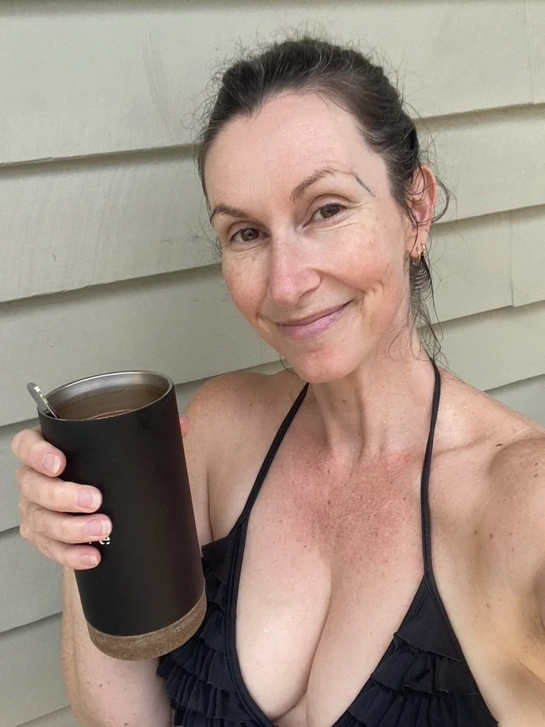 Human Design reader Fiona Lindberg with dark hair in a ponytail smiles, holding a black tumbler with a beverage, standing against beige siding, wearing a black ruffled swimsuit. She has no makeup on, and looks disheveled and happy.