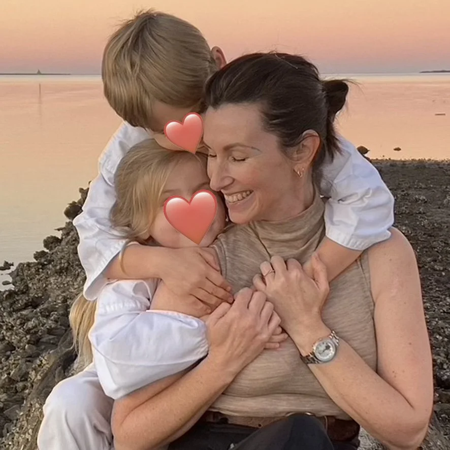 Human Design reader Fiona Lindberg with her two children embracing and smiling with a sunset over water in the background. Two children are kissing her cheek, and the Fiona looks joyful.