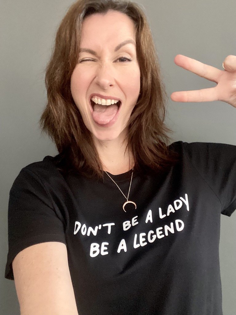Human Design reader Fiona Lindberg with brown hair making a peace sign and winking, wearing a black T-shirt with white text which reads 'Don't be a lady, be a legend', against a grey background.