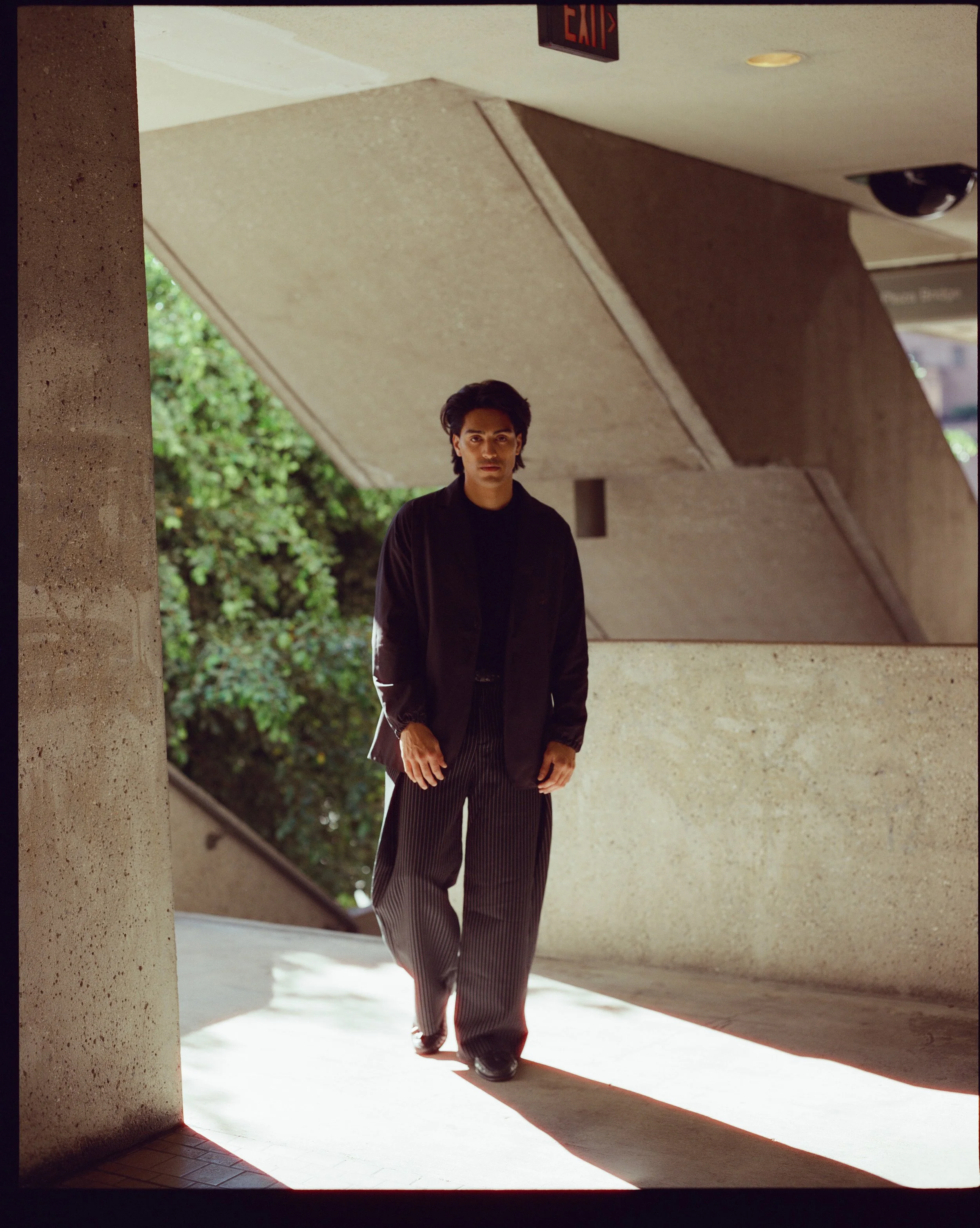 A young man with dark hair dressed in a black jacket and striped pants walking out from underneath a concrete overhang near a building, with an exit sign visible above.