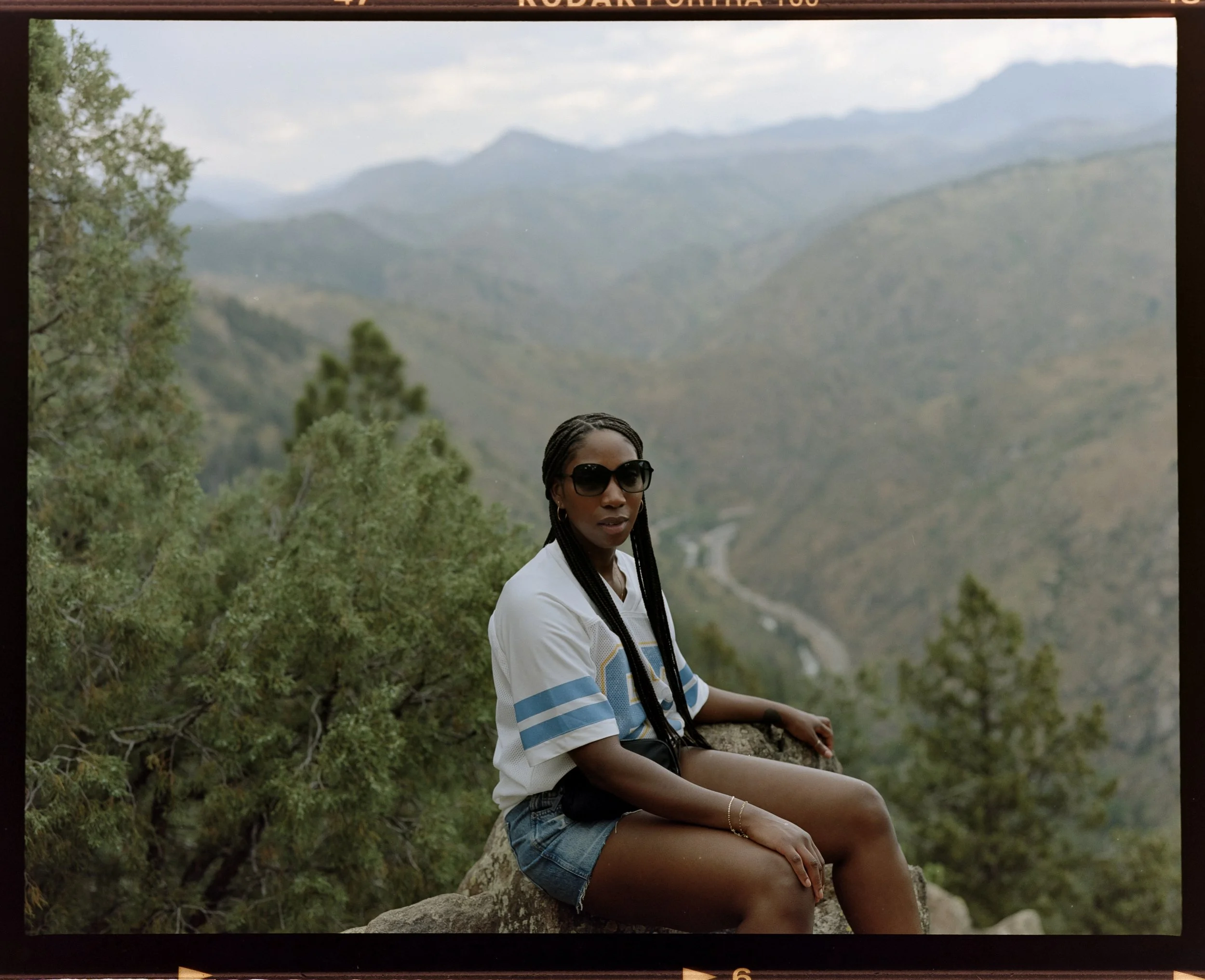 A woman with braided hair and sunglasses sitting on a rock in a scenic mountainous outdoor setting with lush greenery and cloudy sky.