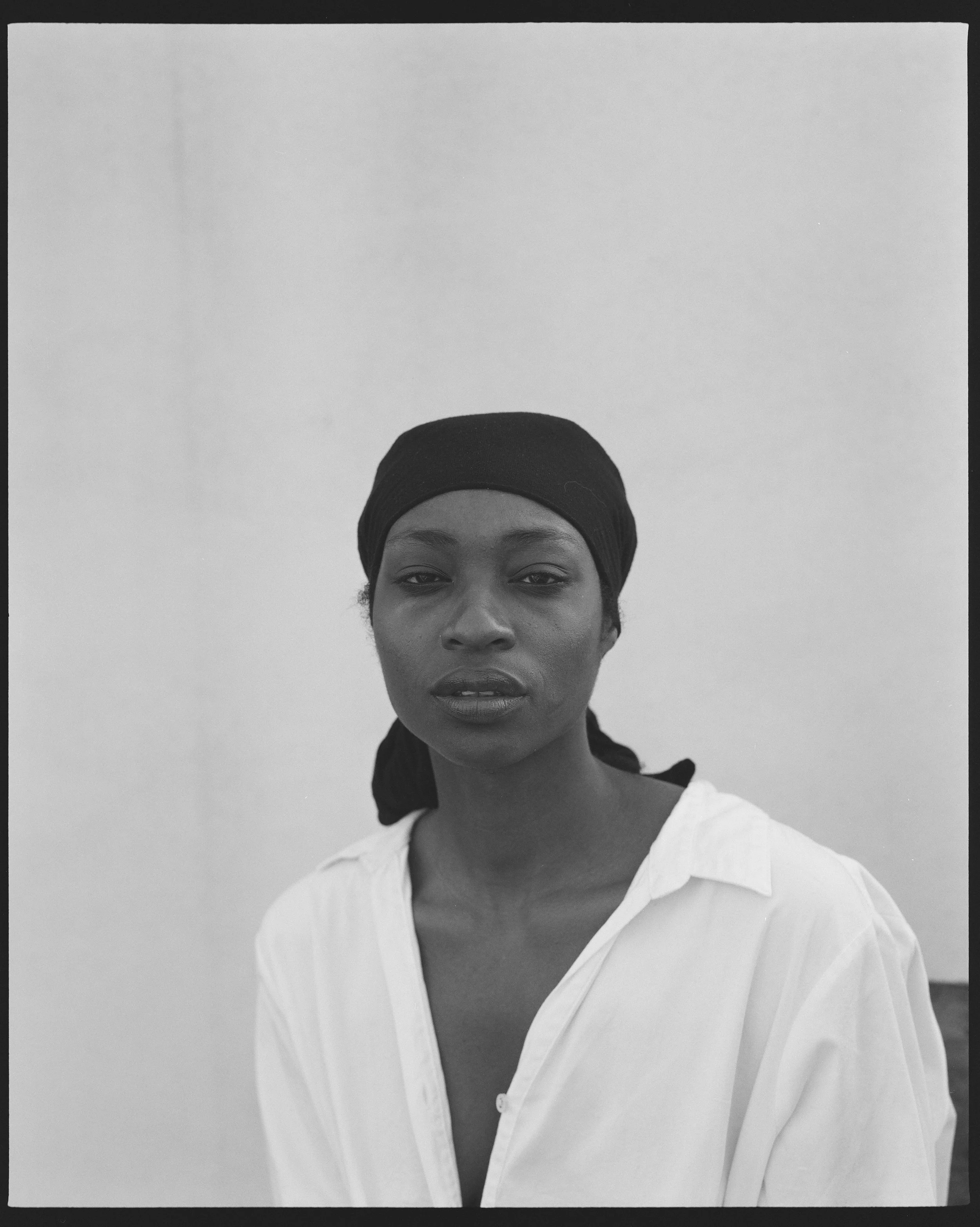 A black and white portrait of a woman with dark eyes and full lips, wearing a headwrap and a white shirt with an open collar. The background is plain and light-colored.