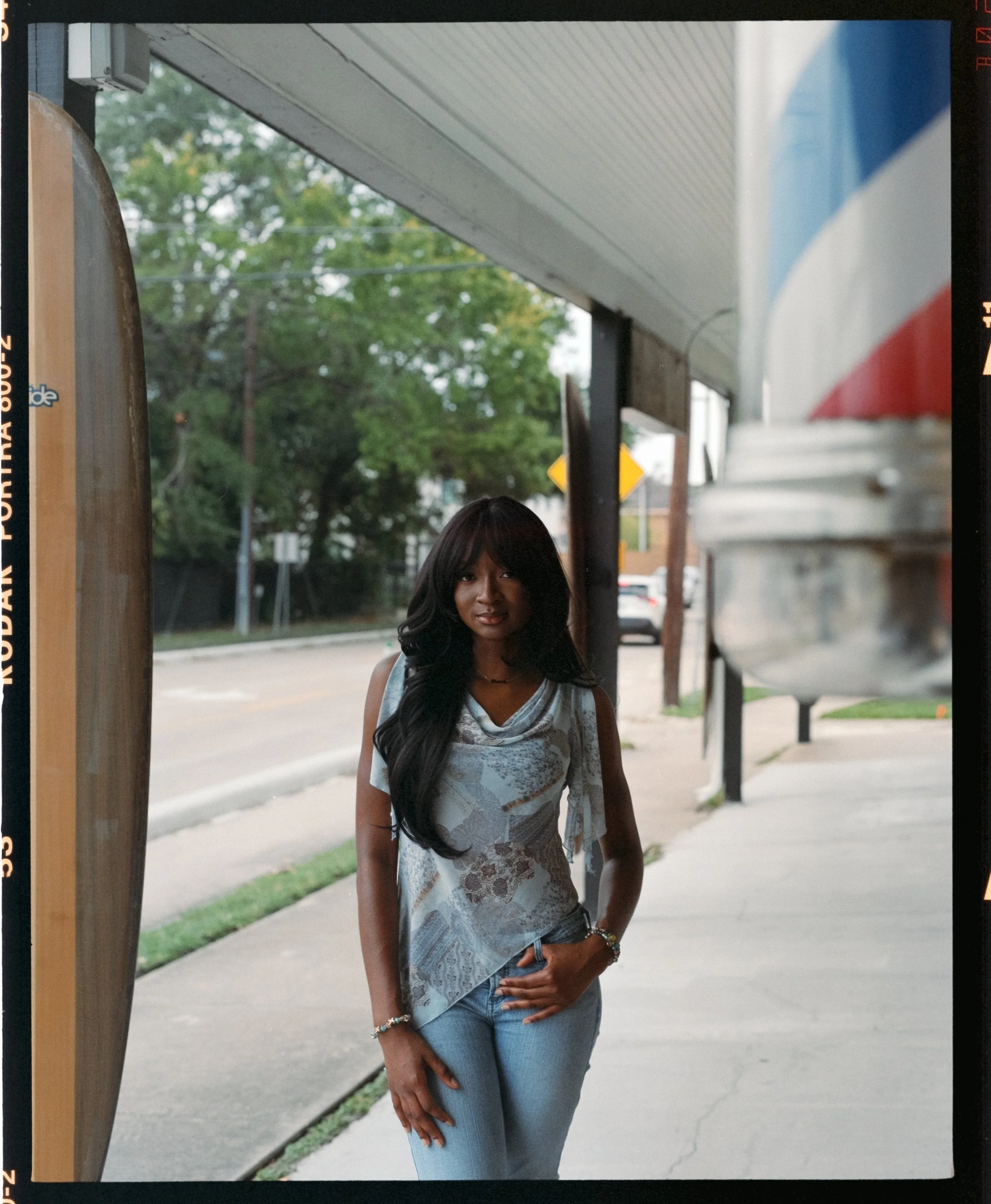 A woman with long dark hair standing on a sidewalk outdoors near a building and trees in the background, wearing a sleeveless top, jeans, and jewelry.