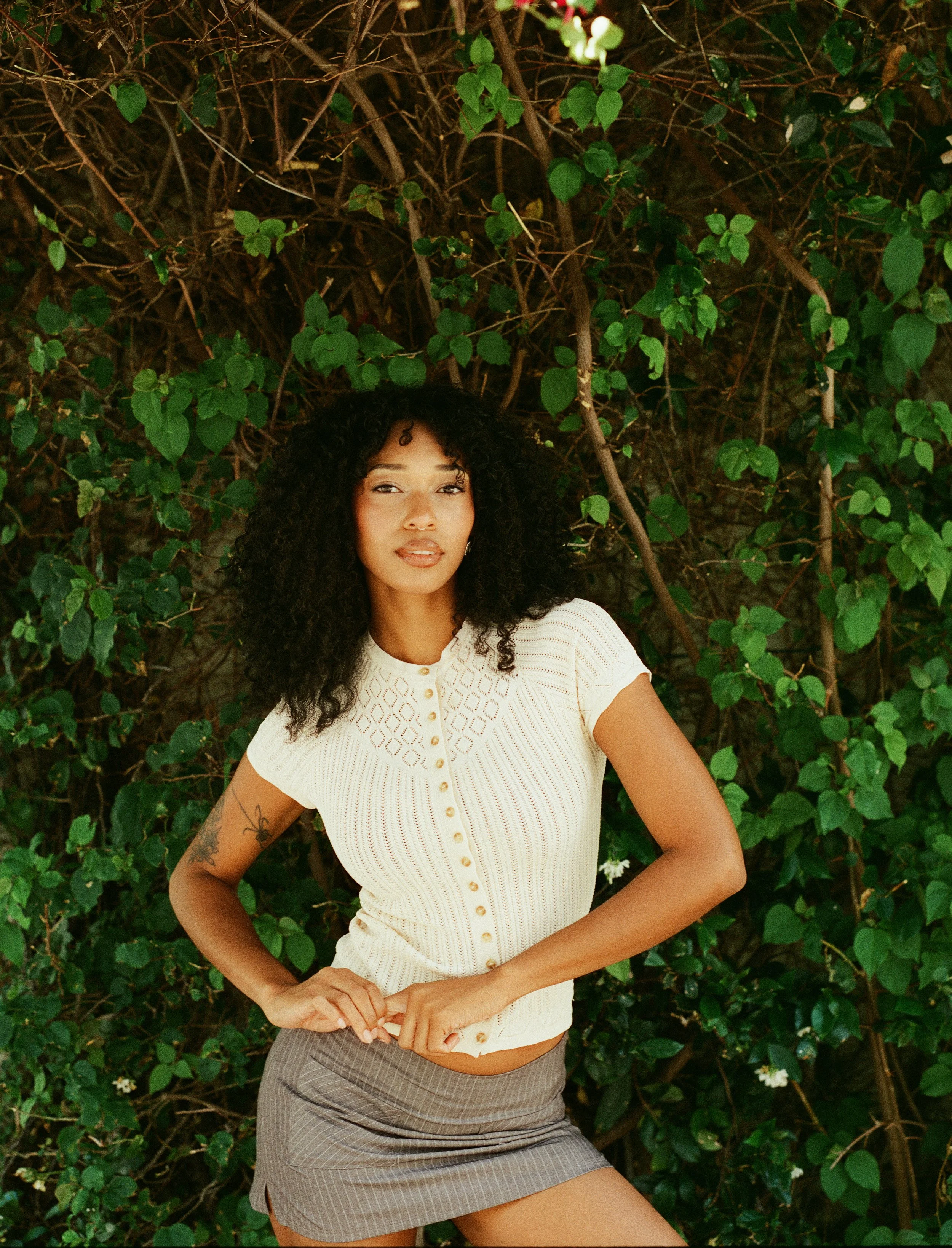 A woman with curly dark hair wearing a beige button-up short sleeve top and a gray skirt is standing outdoors, posing in front of a dense background of green leaves and vines.