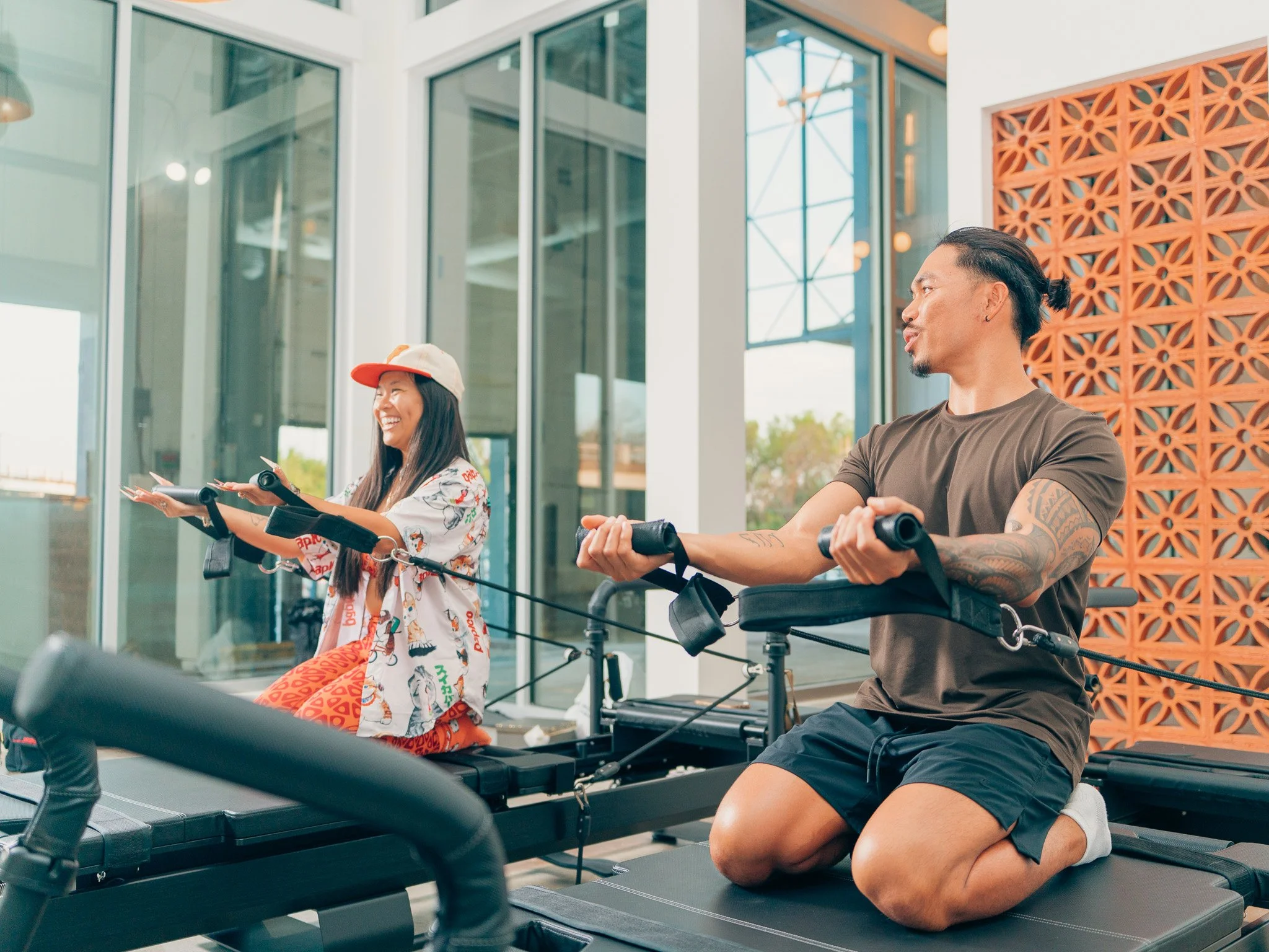Two people, a woman and a man, are exercising on rowing machines indoors. The woman is smiling, wearing a colorful outfit with a baseball cap, and the man is focused, with tattoos, wearing a brown t-shirt and black shorts.