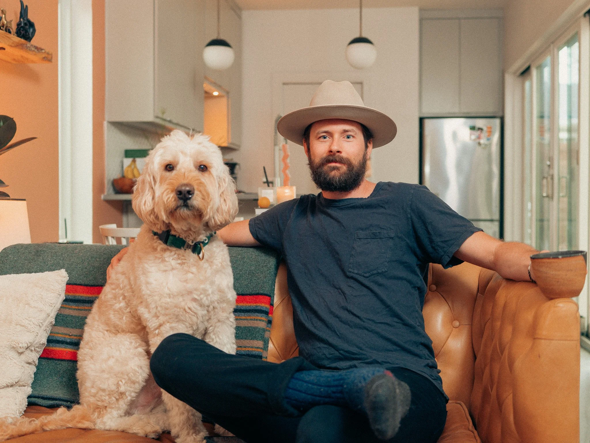 A man with a beard and wearing a wide-brimmed hat and a dark T-shirt is sitting on a tan couch with a fluffy dog sitting beside him in a cozy living room.