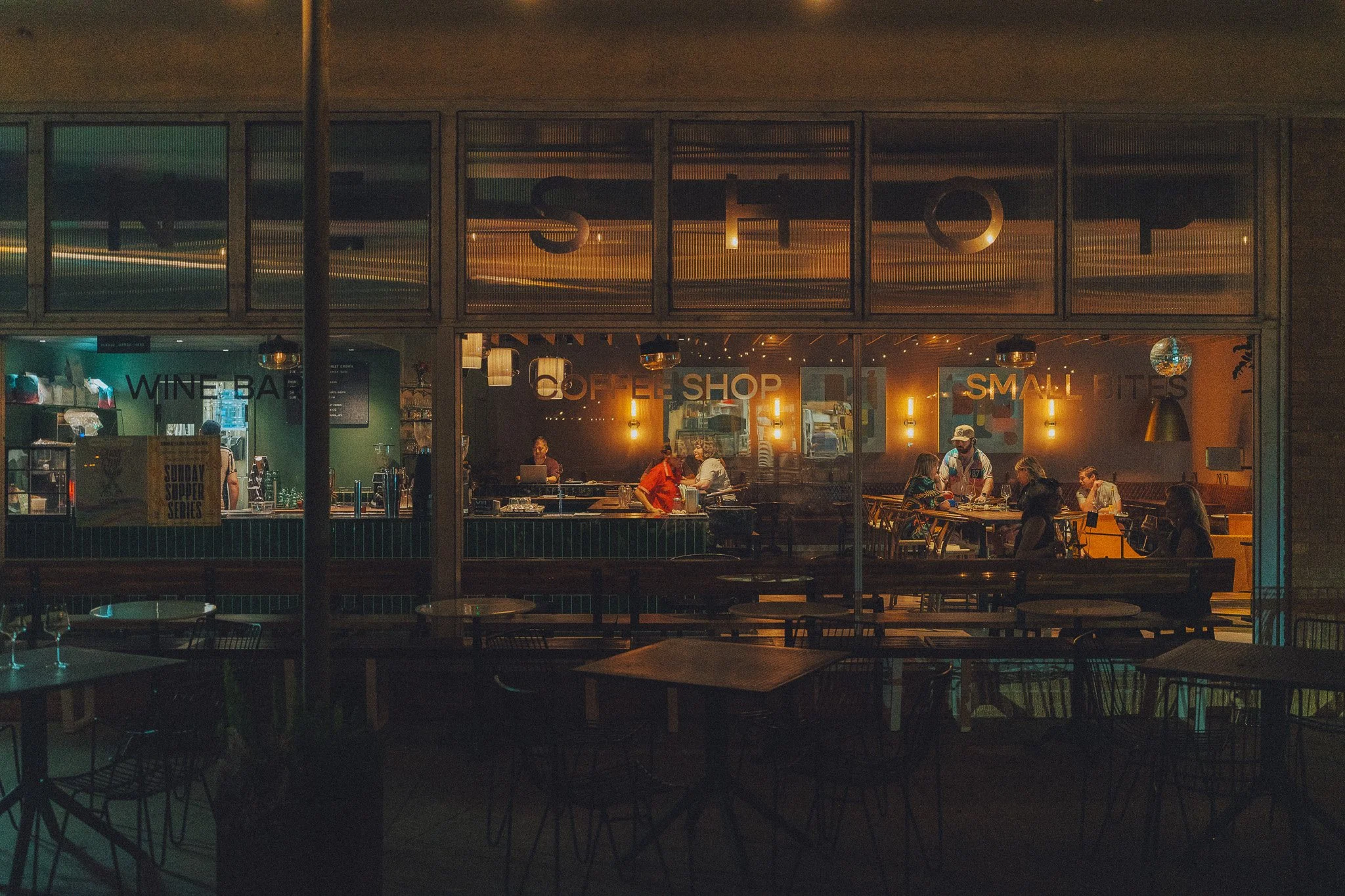 Inside view of a cozy restaurant during the evening, with customers dining and staff behind the counter. The warm lighting creates an inviting atmosphere, with signs for wine bar, coffee shop, and small bites visible.