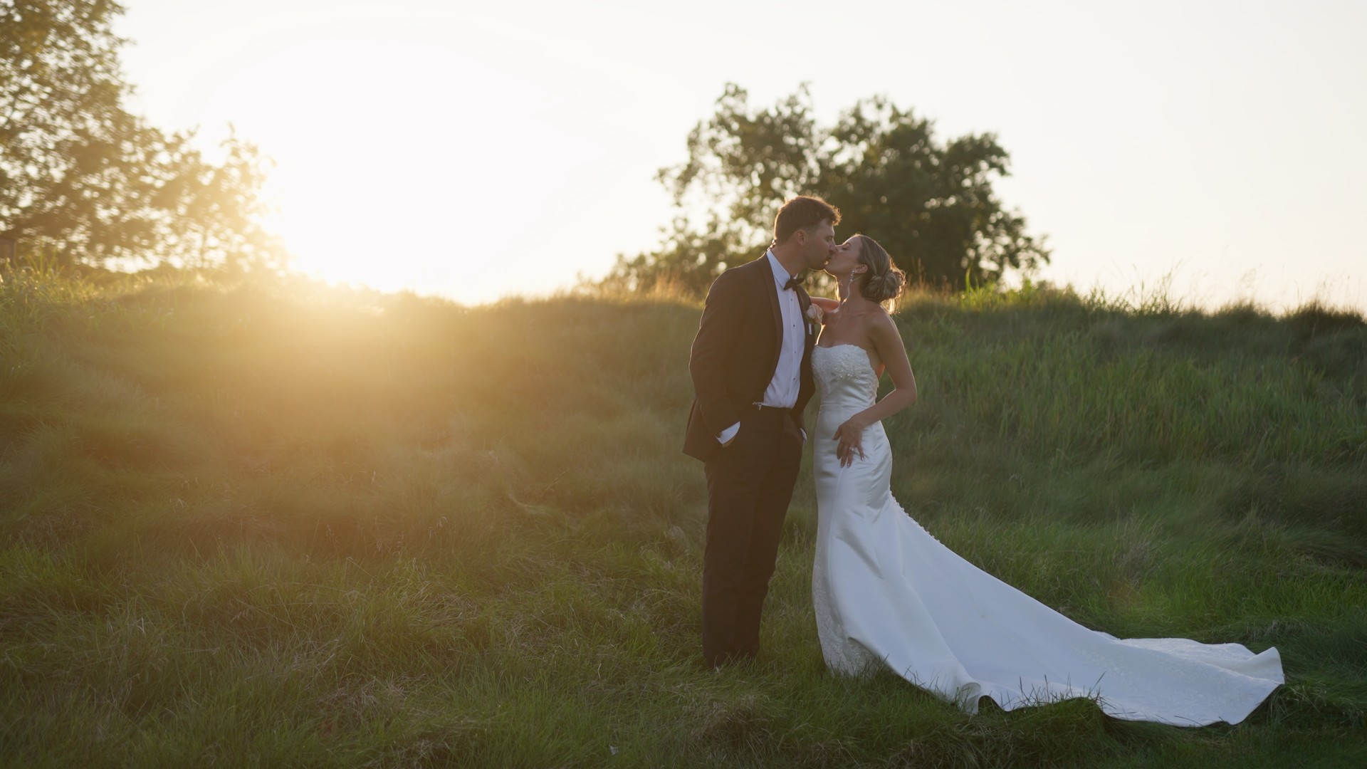 A bride and groom kissing outdoors during sunset, standing on grass with trees in the background.