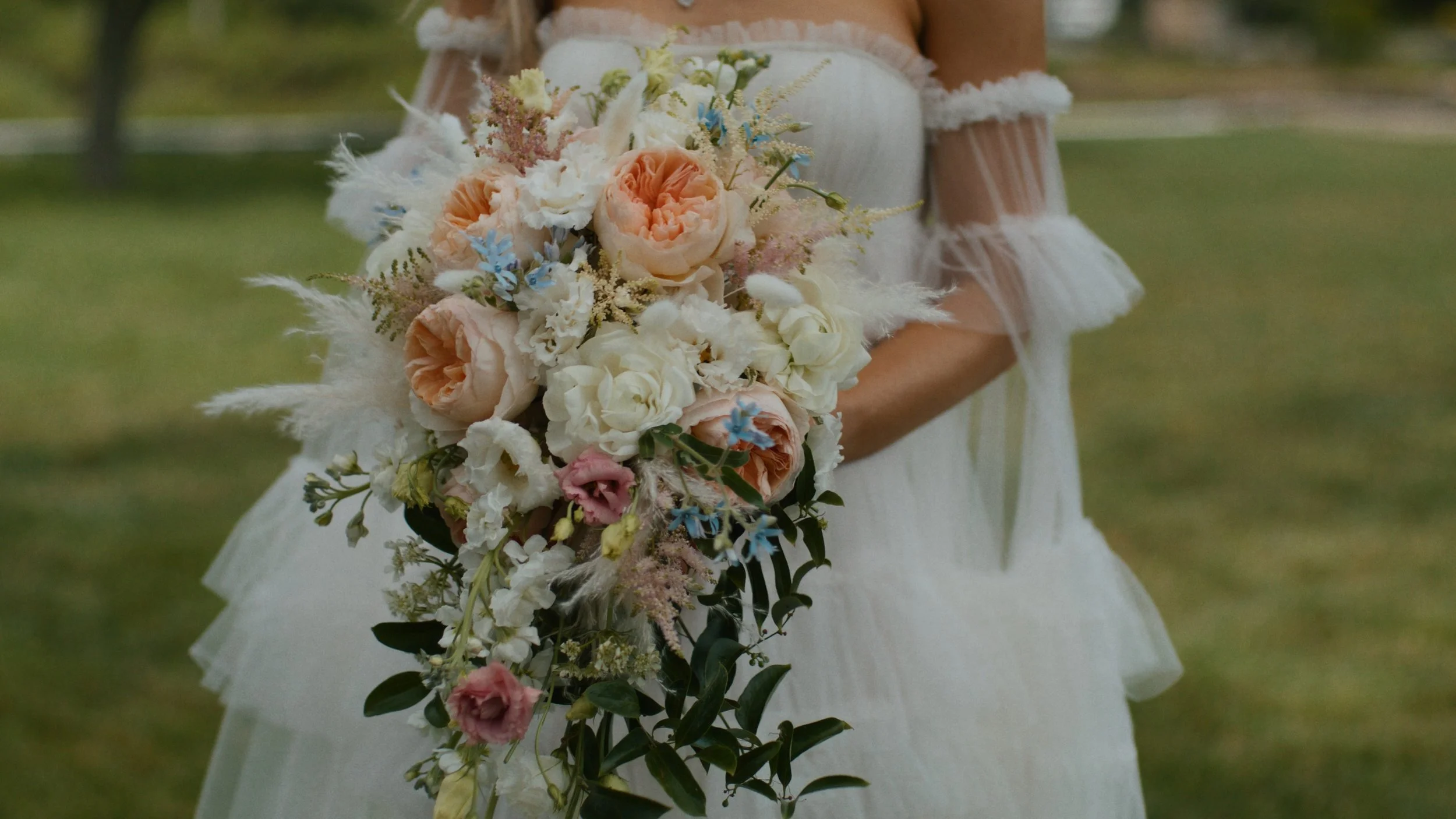 A bride holding a cascading bouquet of peach, white, pink, and blue flowers, with greenery, in an outdoor setting.