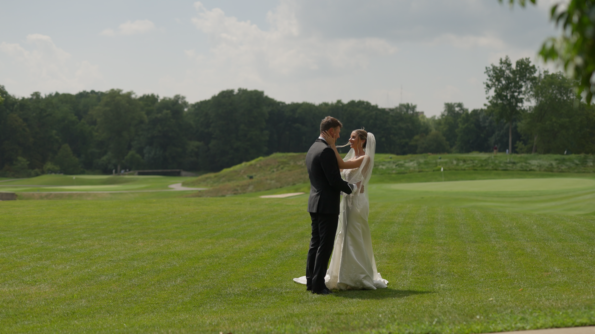 A bride and groom standing closely together on a lush green golf course, sharing a moment under a slightly cloudy sky with trees in the background