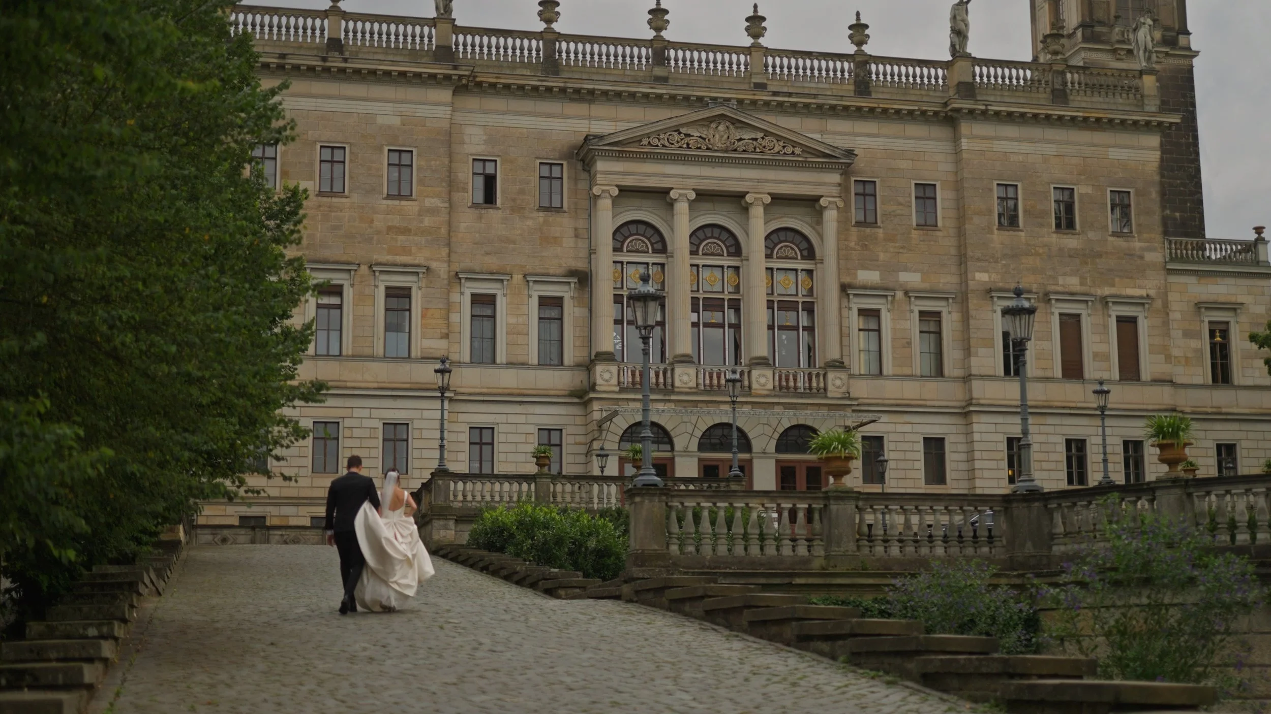 A bride and groom walking up outdoor stone stairs toward a large, historic building with ornate windows, columns, and statues, surrounded by greenery and lampposts.