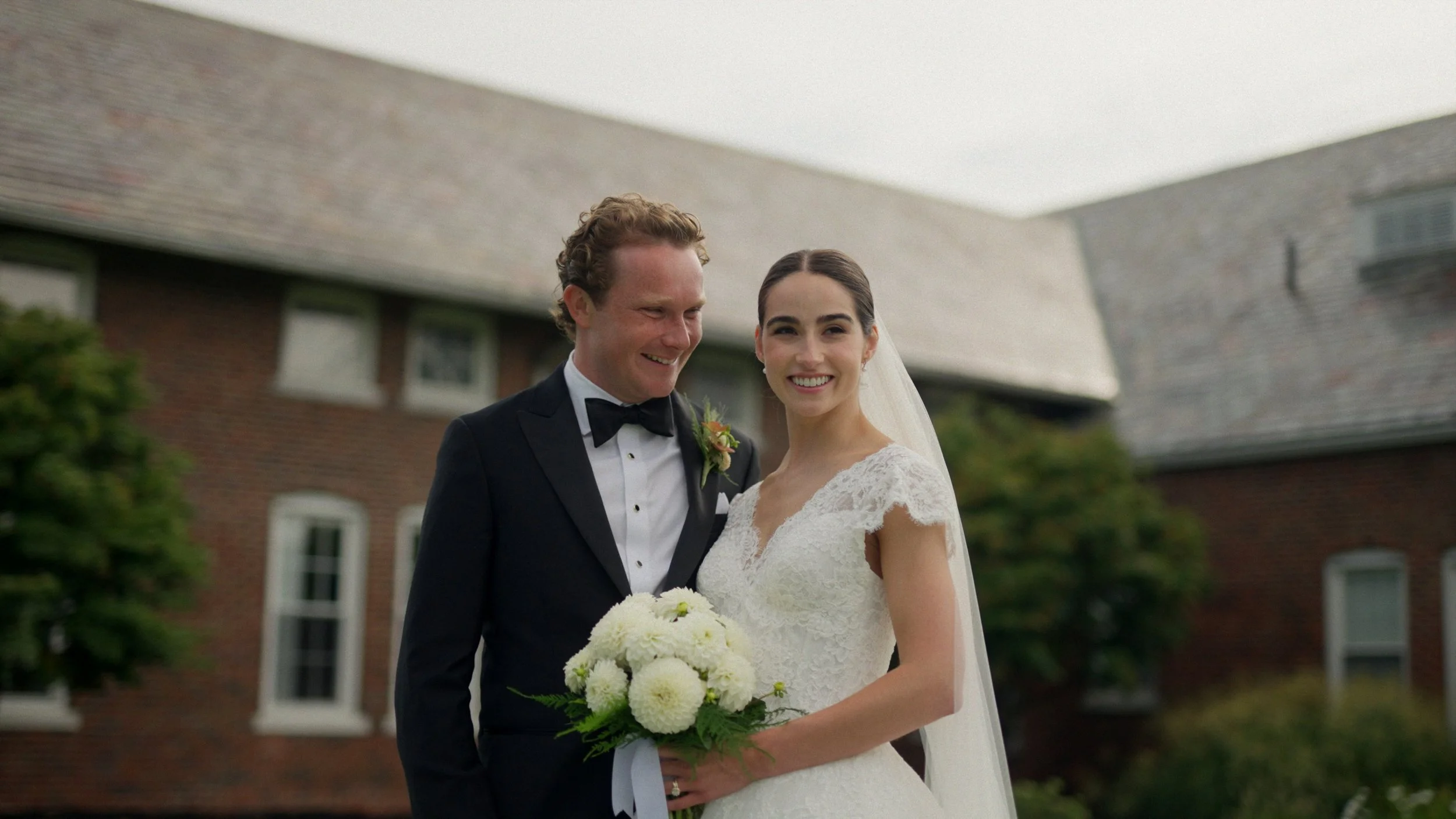 A bride and groom standing together outdoors in front of a brick building with windows, smiling and dressed in wedding attire. The groom is wearing a black tuxedo with a bow tie, and the bride is in a white lace wedding gown holding a bouquet of white flowers.