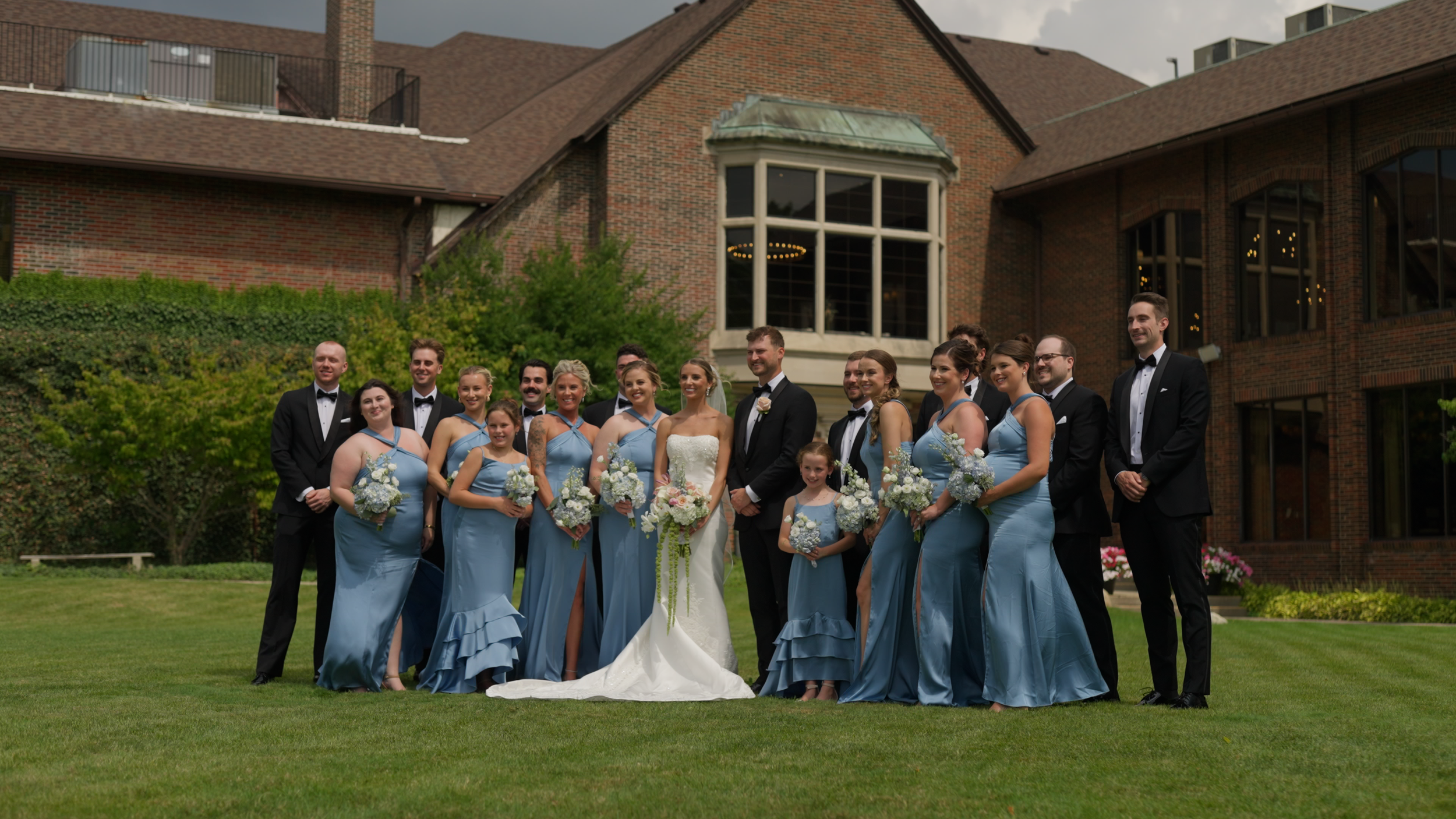 A wedding party posed outdoors in front of a brick building with large windows. The group includes the bride in a white gown, groom, bridesmaids in matching light blue dresses, and groomsmen in black tuxedos. The bridesmaids hold bouquets of white flowers, and the bride holds a cascading bouquet. The group is smiling and standing on a grassy area.
