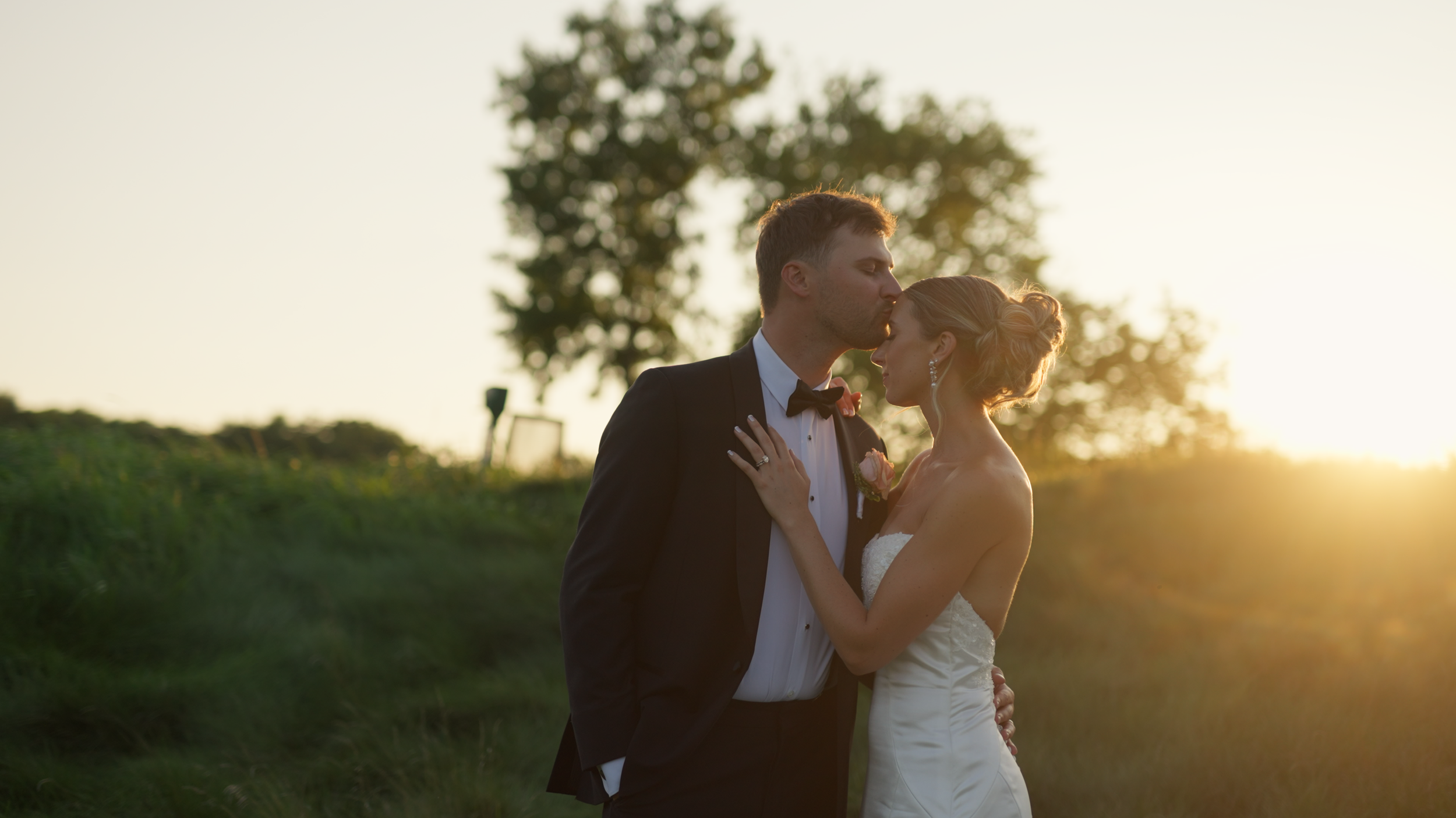 A bride and groom sharing a kiss outdoors during sunset, with trees in the background.