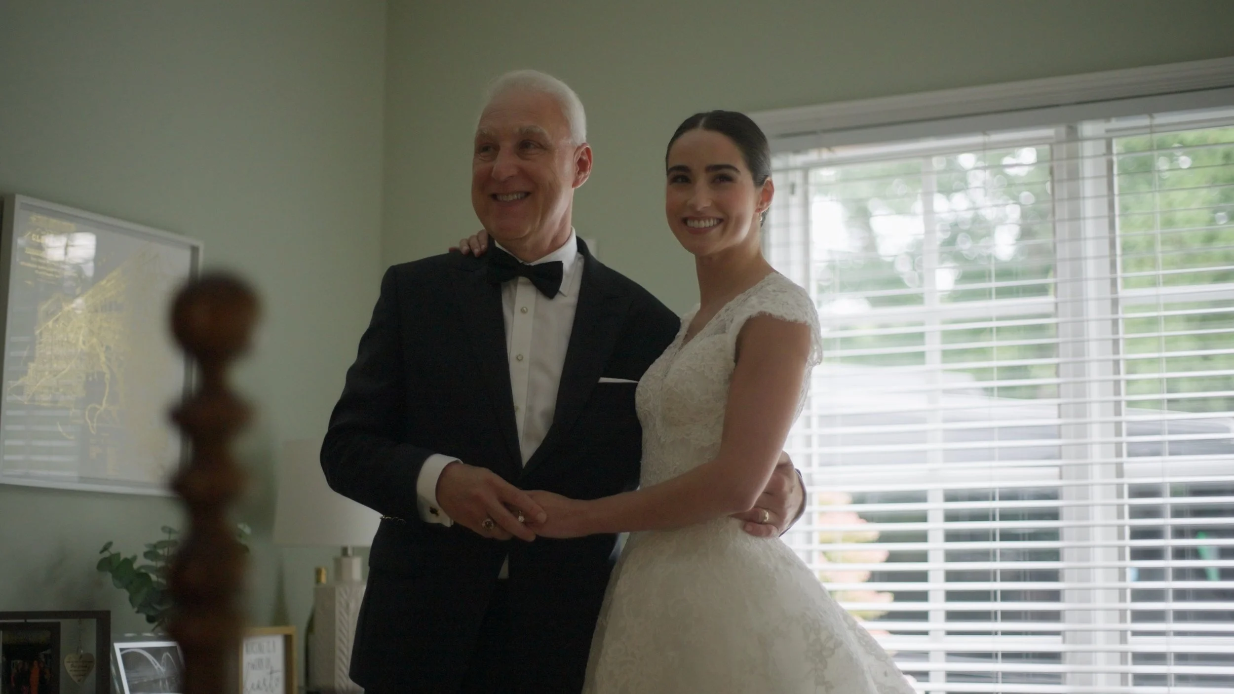 A man and woman in wedding attire smiling and embracing indoors near a window with blinds.