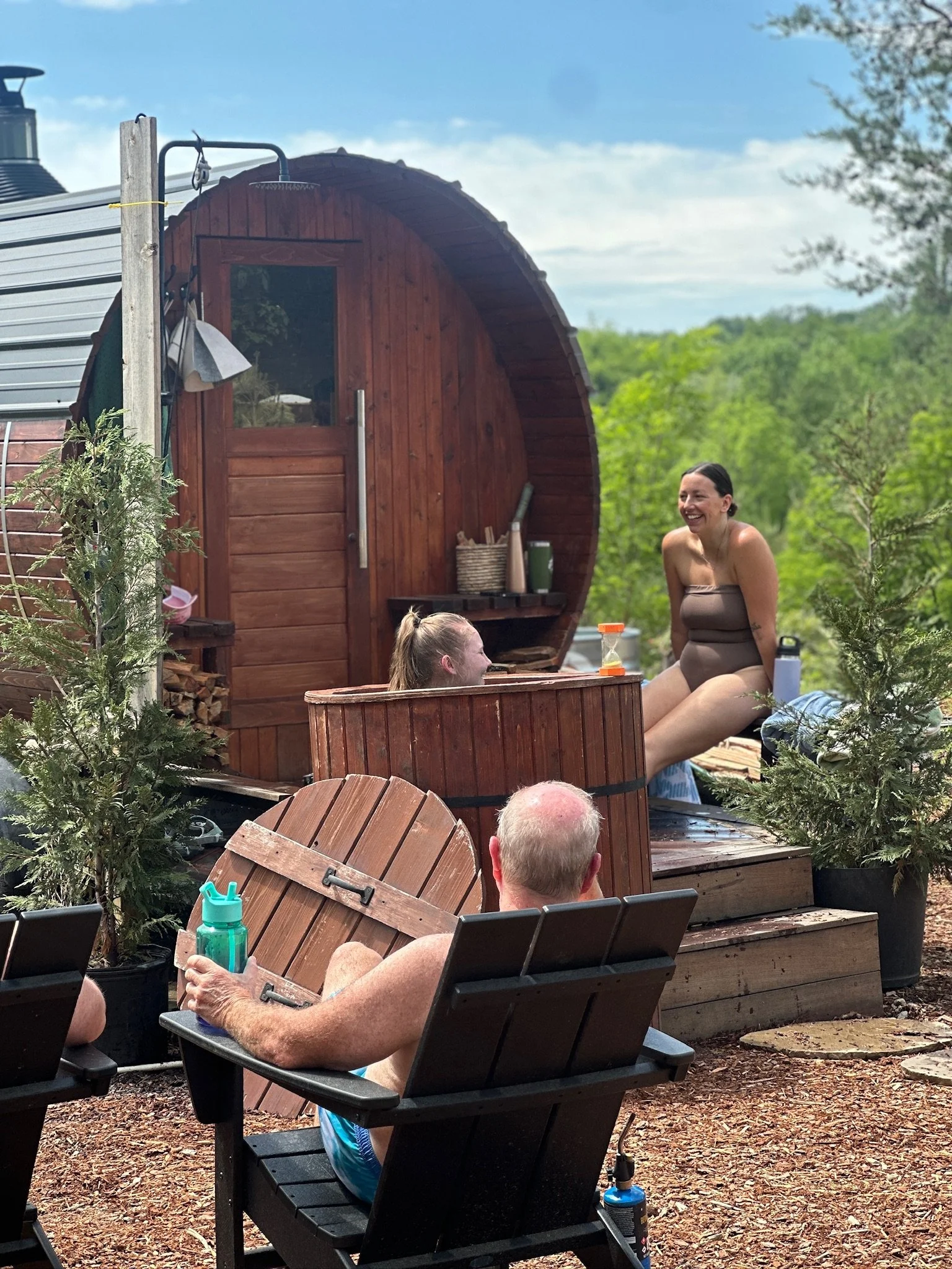 People enjoying outdoor hot tub and sauna on a sunny day in a green, wooded backyard.